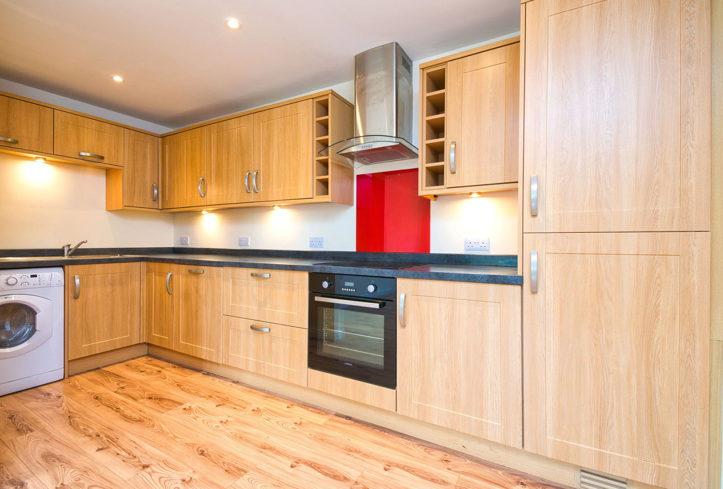 Modern kitchen with wooden cabinets, black countertop, built-in oven, stainless steel range hood, washing machine, and red accent wall.
