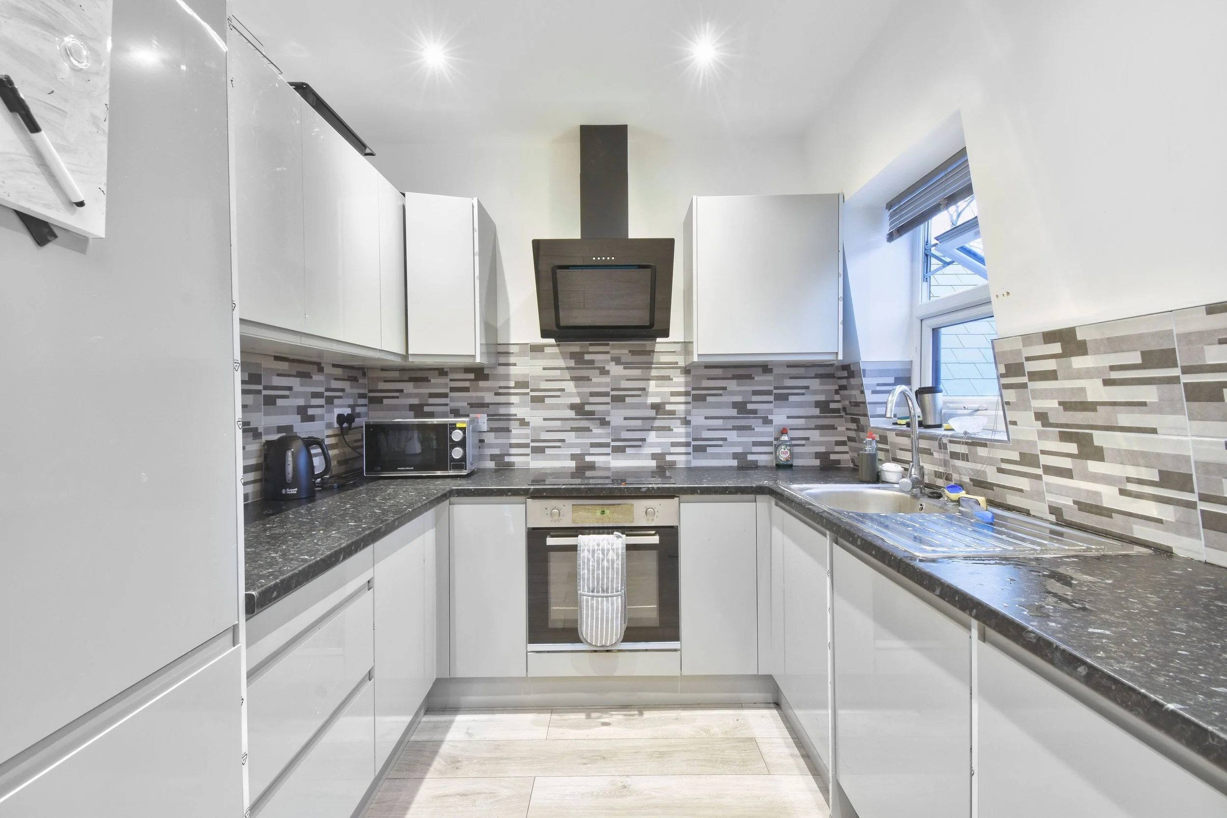 Modern kitchen with white cabinets, black granite countertops, stainless steel appliances including an oven, microwave, kettle, and toaster, a tile backsplash, a window above the sink, and bright ceiling lights.
