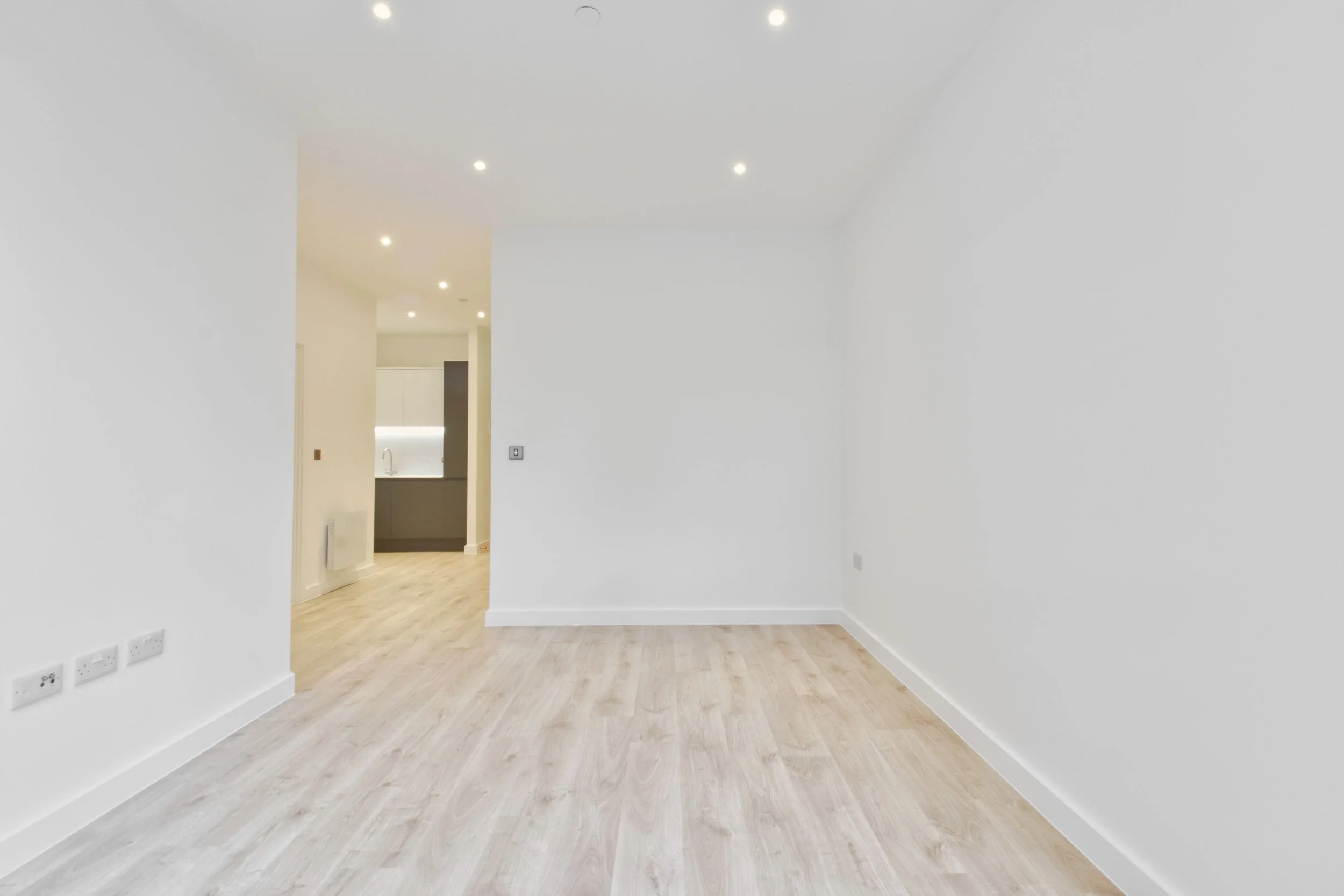 Empty white room with light-colored wooden flooring, multiple electrical outlets on the wall, and a doorway leading to a kitchen area with a sink and cabinetry.