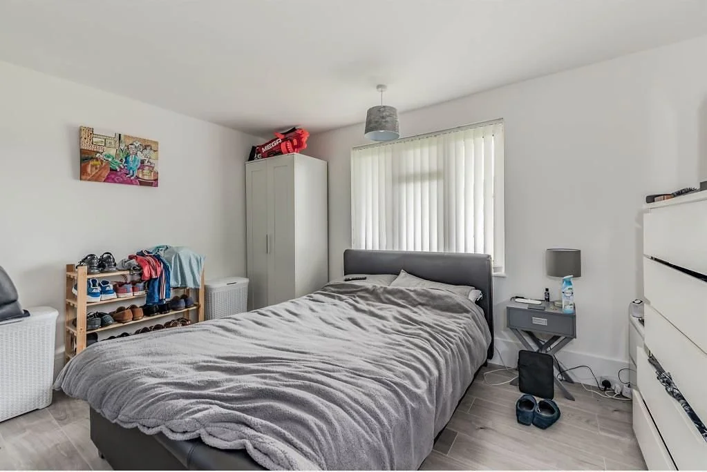 A bedroom with a bed, a window with blinds, a small bedside table with a lamp and water bottle, a white dresser, a wardrobe, a shoe rack, and casual clothing and shoes on the floor.