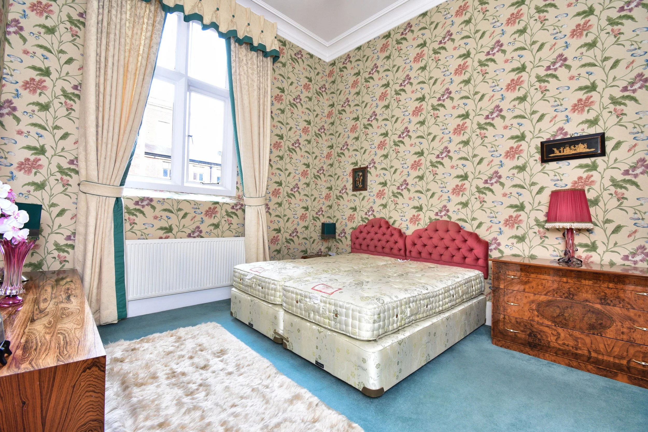 A bedroom with floral wallpaper, a window with curtains, a pink tufted headboard, a mattress without sheets, a wooden nightstand with a red lampshade, a fluffy beige rug, and a set of small framed pictures on the wall.