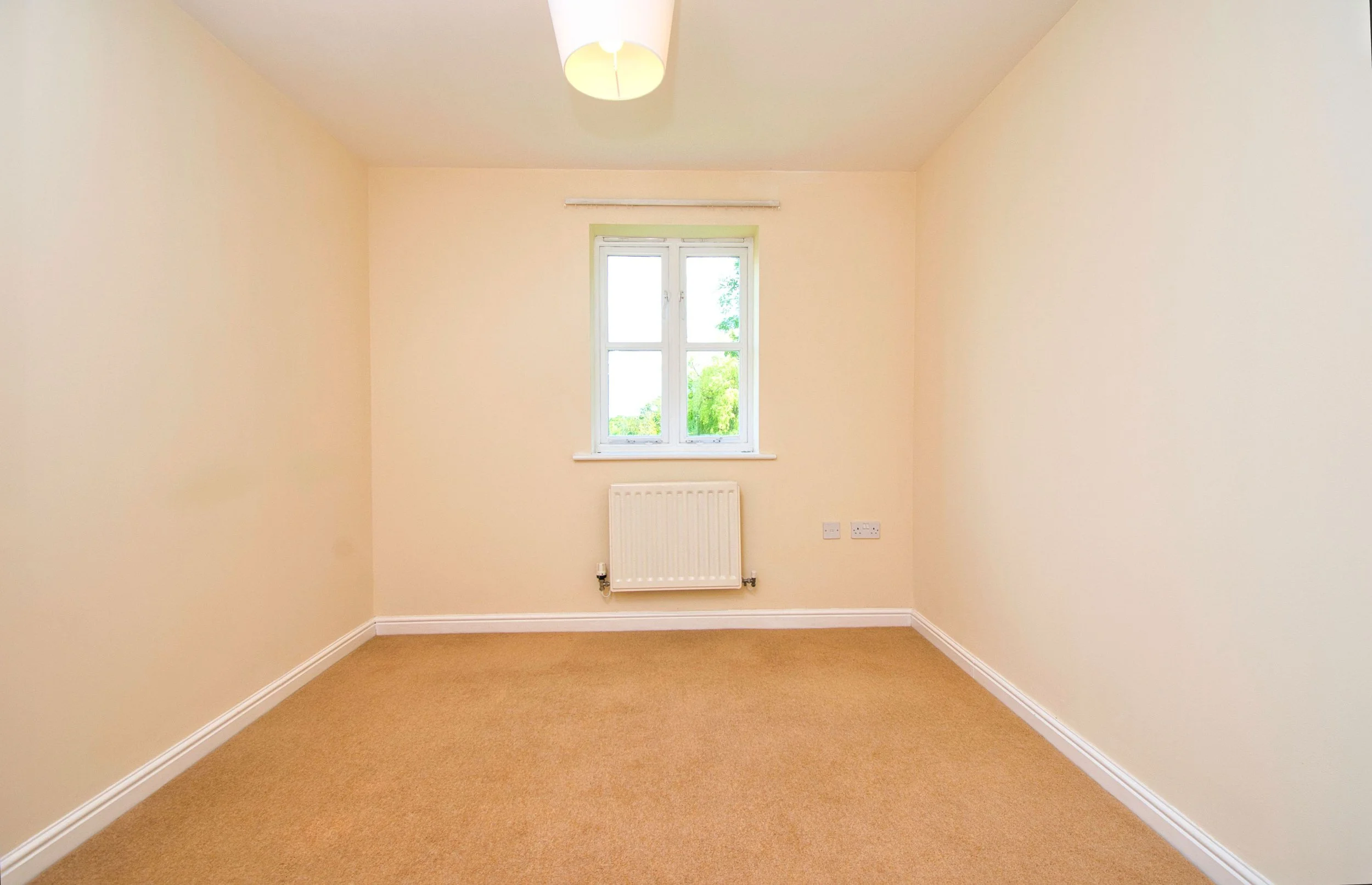 Empty beige carpeted room with cream-colored walls, white trim, a small window with a view of trees, a white radiator underneath the window, and an overhead light fixture.