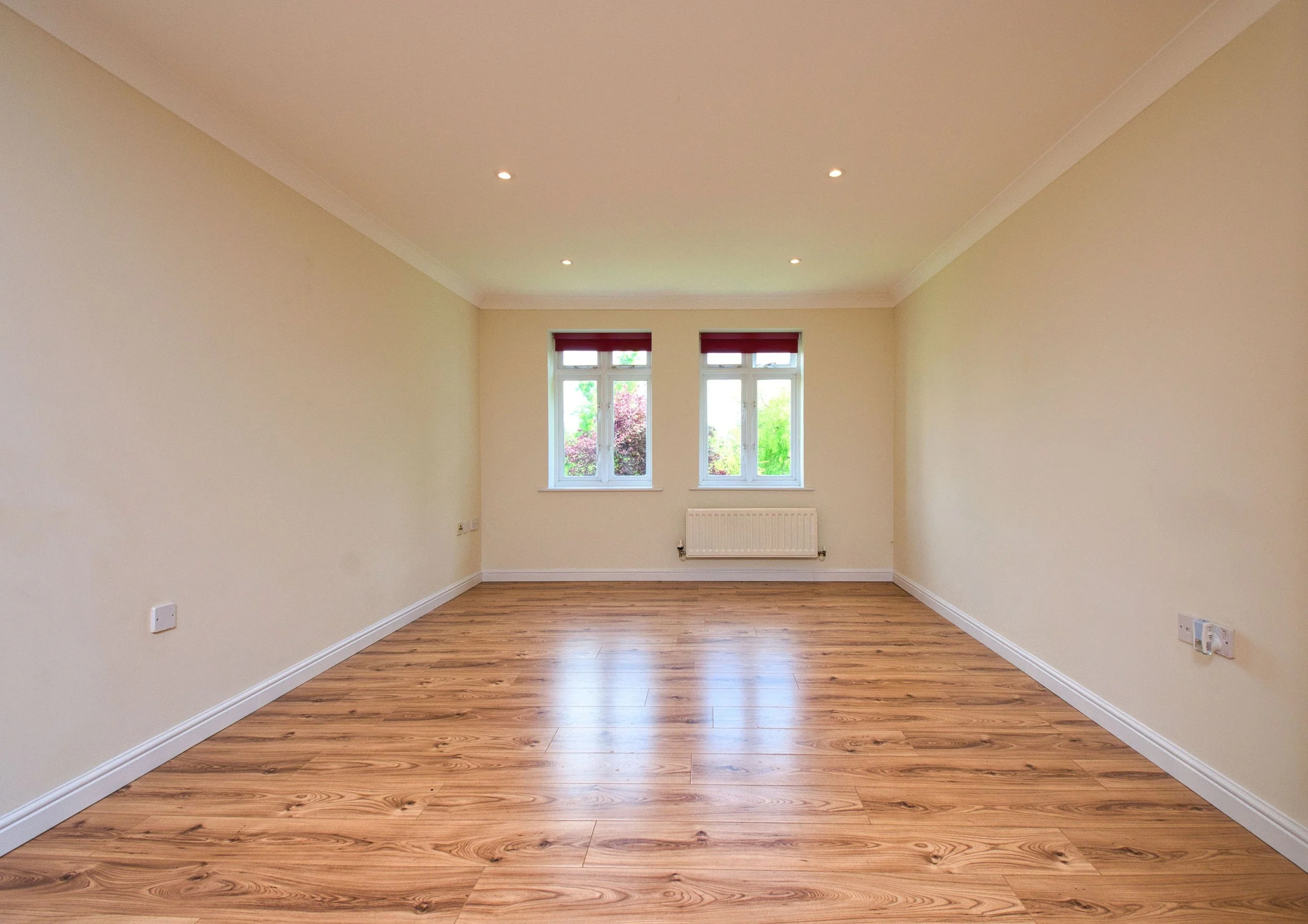 Empty living room with beige walls, wooden flooring, three windows with red blinds, white radiator, and ceiling-mounted lights.