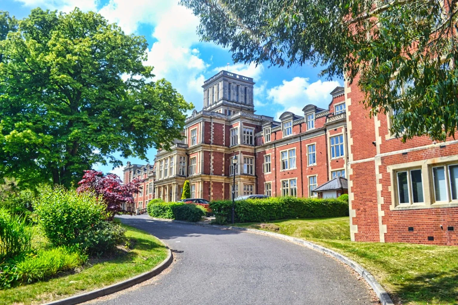 A brick apartment building with multiple windows, surrounded by green trees, bushes, and a curved paved road.