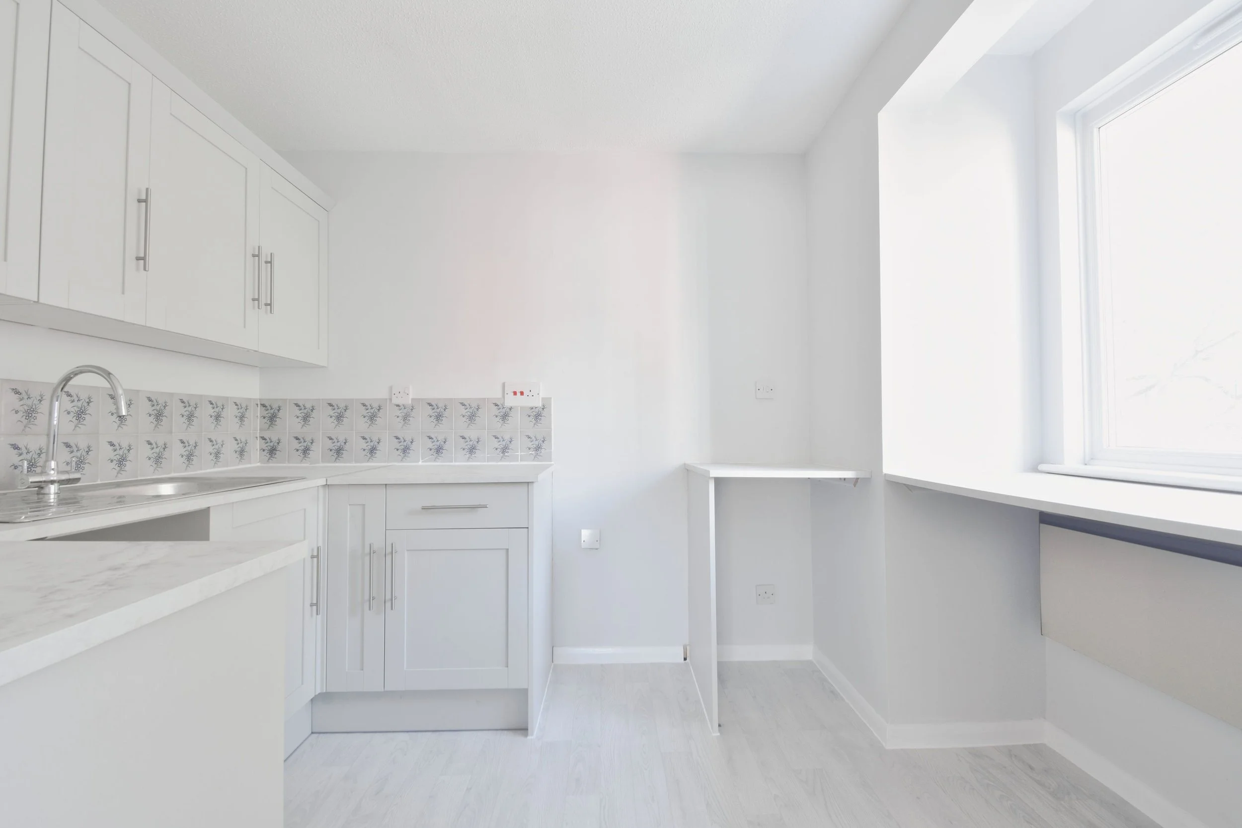 Empty white kitchen with cabinets, sink, window, and light-colored flooring.