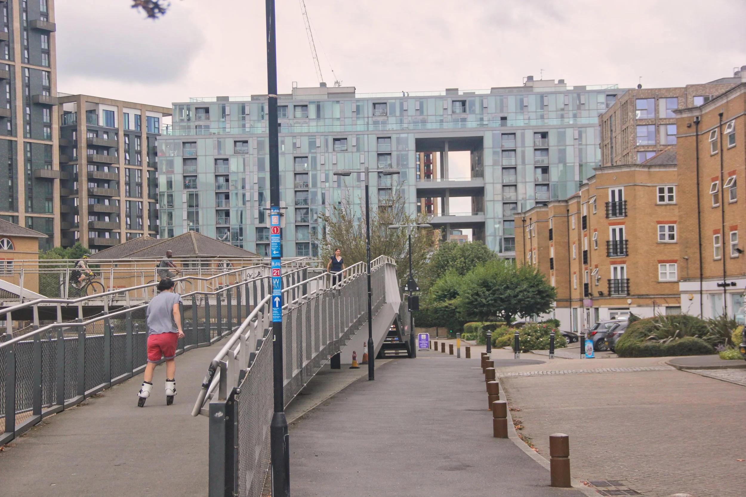 Urban scene with a pedestrian bridge, people skating, city buildings, and trees.