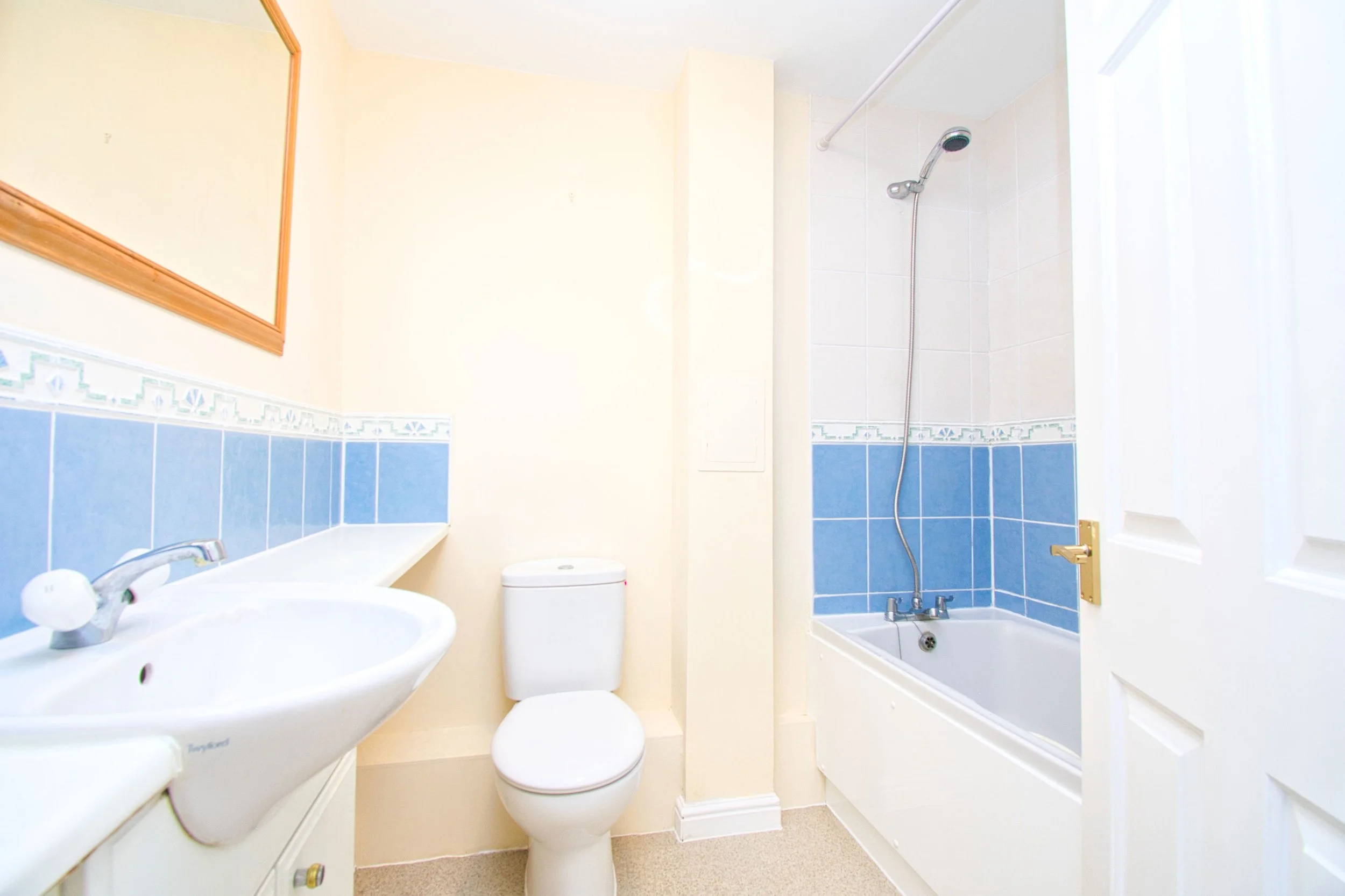 Small bathroom with a white sink, toilet, and bathtub with a showerhead. The walls are painted light yellow with a decorative blue tile border running halfway up. A mirror with a wooden frame hangs on the wall above the sink, and the bathroom door is