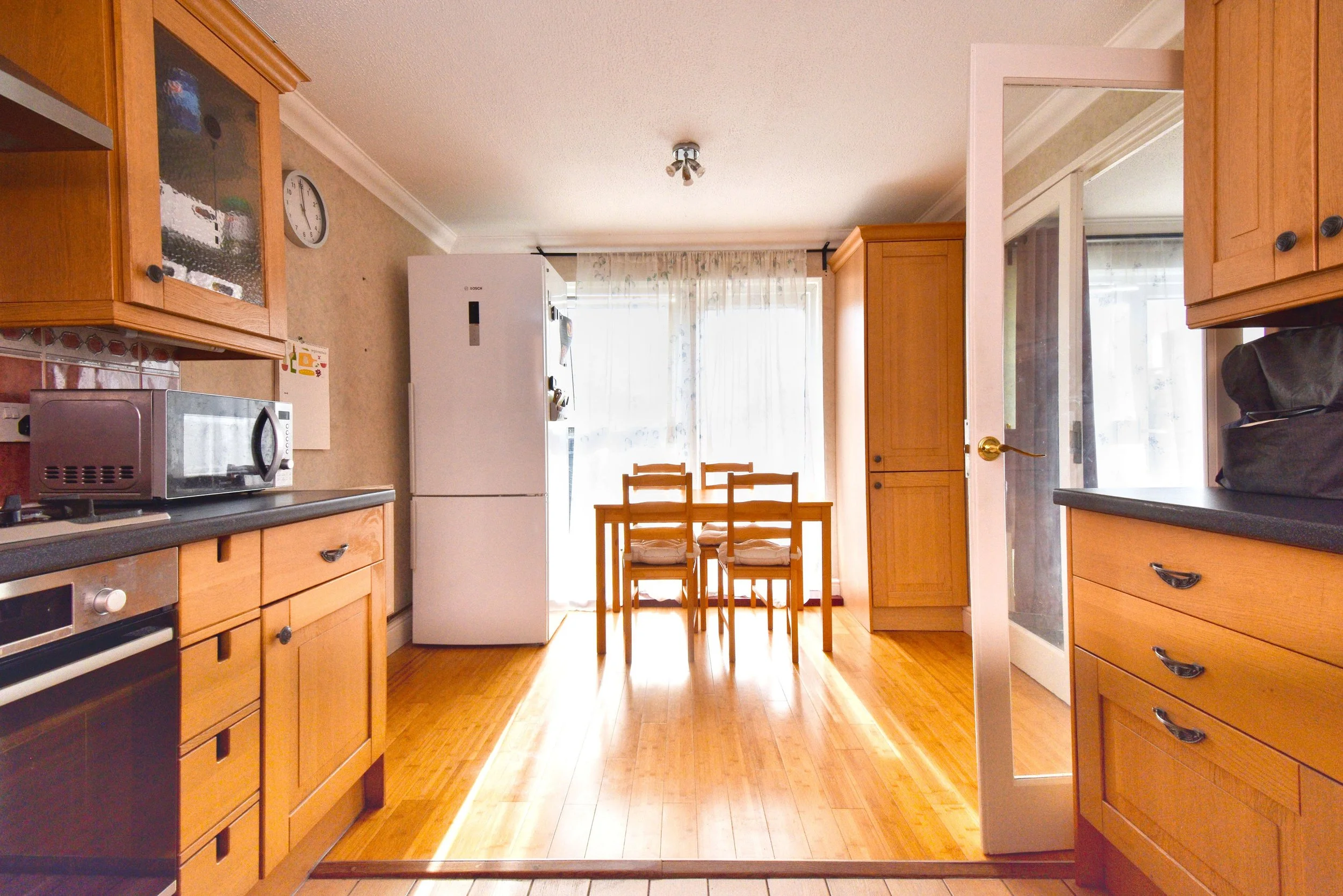A kitchen view showing wooden cabinets, a white refrigerator, a microwave, and a dining table with chairs near a door leading to a room with a window, curtains, and sunlight.