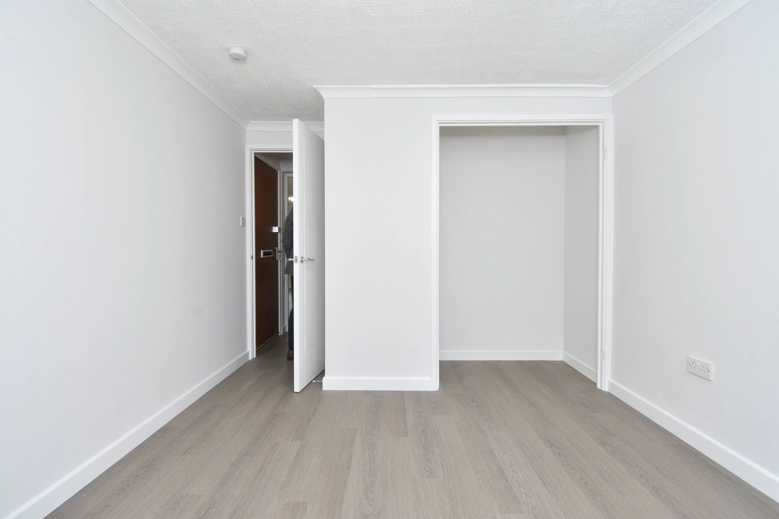 Empty room with white walls and light-colored wooden flooring, open door revealing a person in the doorway.