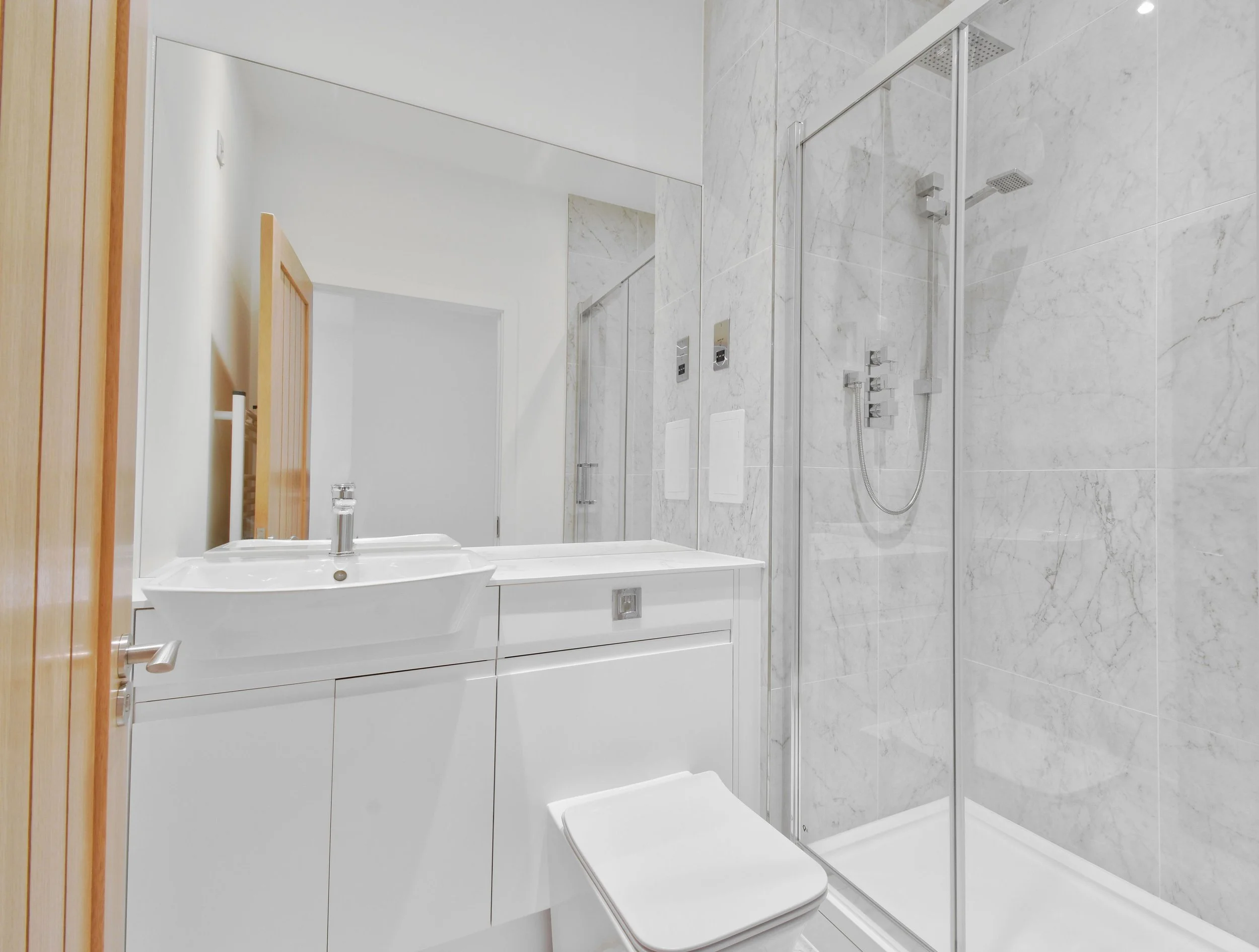 A modern bathroom with marble tiles on the walls and floor, featuring a walk-in shower with a rain showerhead, a white sink with a mirror above, and a toilet.