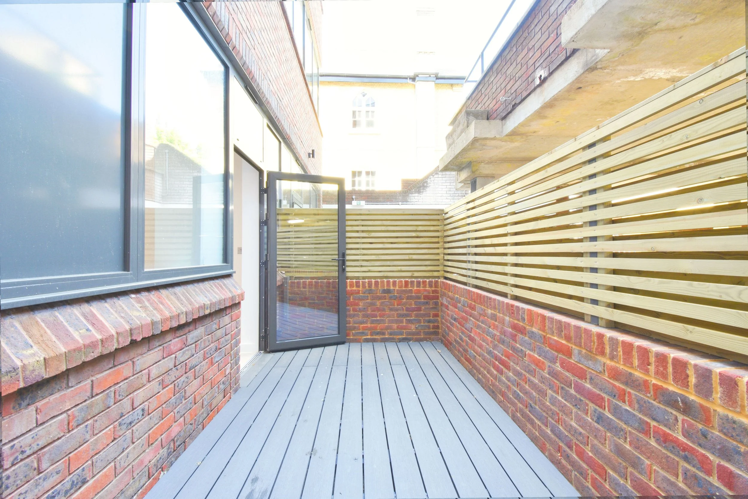 A small balcony or outdoor space with red brick walls, gray wooden deck flooring, and a wooden privacy fence. There is a glass door with a black frame leading inside, and a large window on the left side. An open door is visible beneath the window.