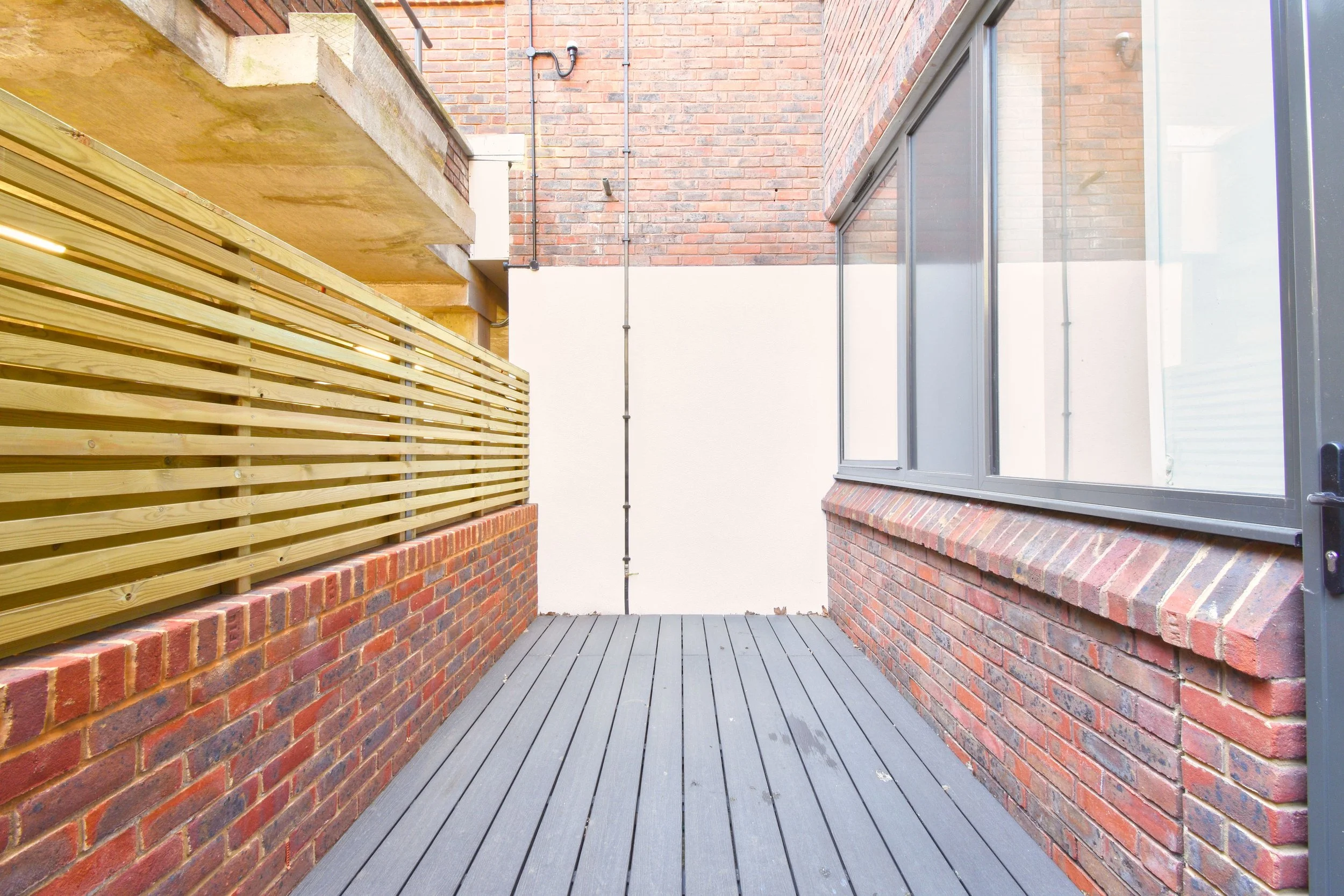 A small brick balcony with a wooden railing, gray wooden deck flooring, a large window, and a brick wall with visible pipes.