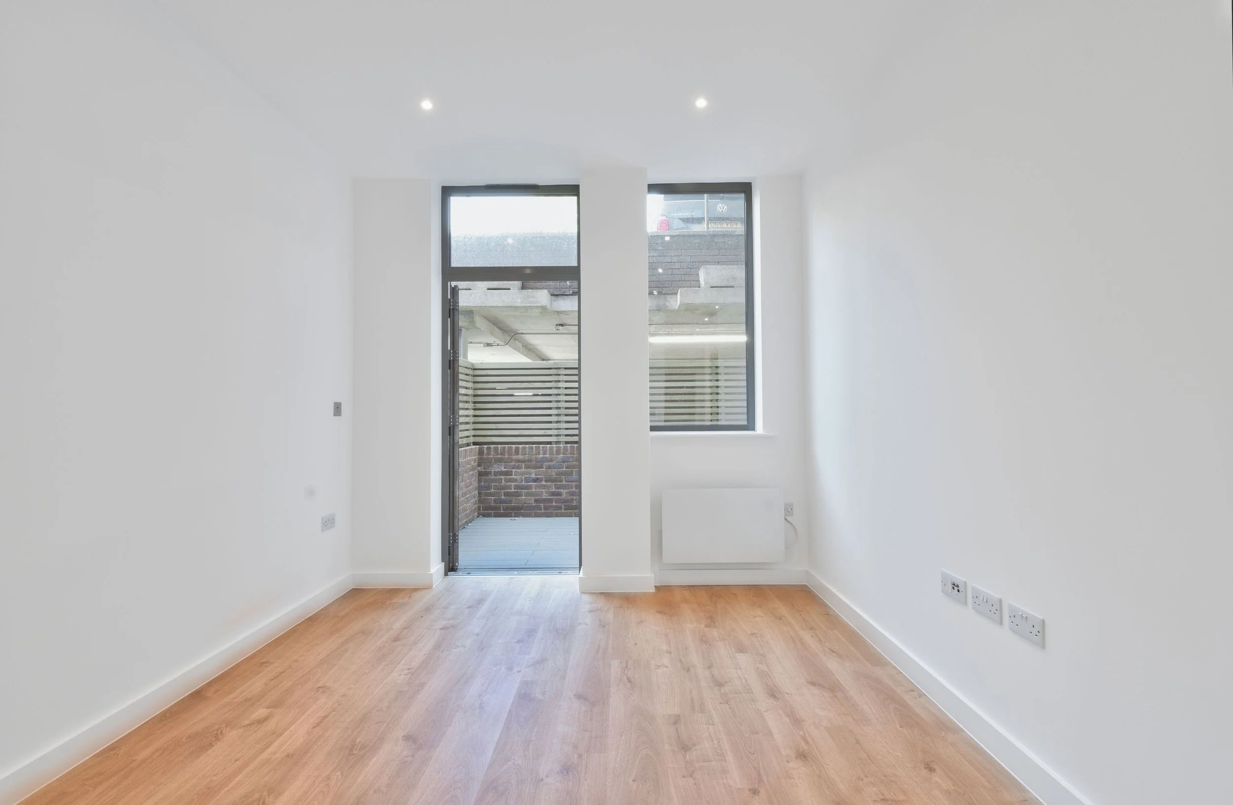 Empty room with white walls, wooden floor, and a large window with a door leading to a small outdoor balcony.
