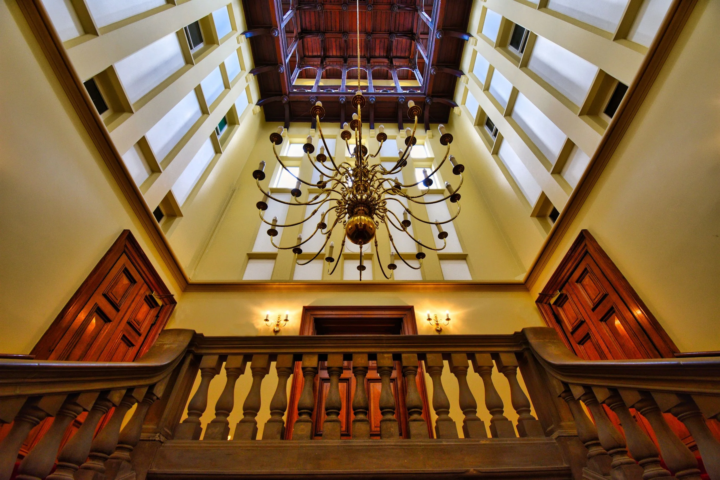 Interior view looking up at a decorative chandelier hanging in a large, multi-story atrium with walls featuring evenly spaced windows and wooden accents.