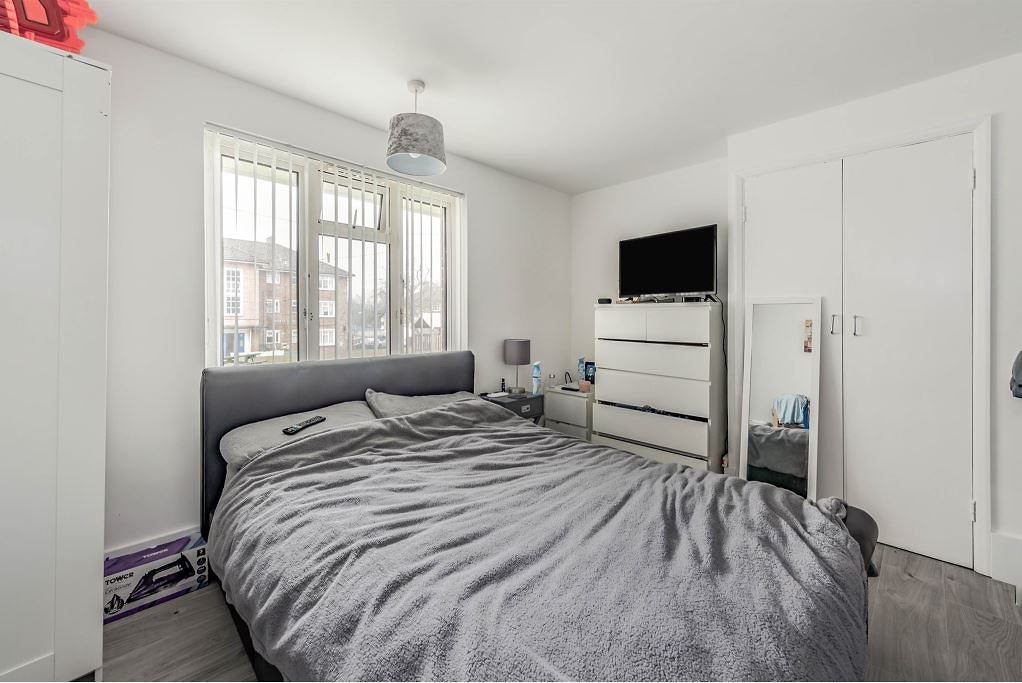 A bedroom with a bed, bedside tables, a dresser with a TV, a mirror, and windows with vertical blinds.