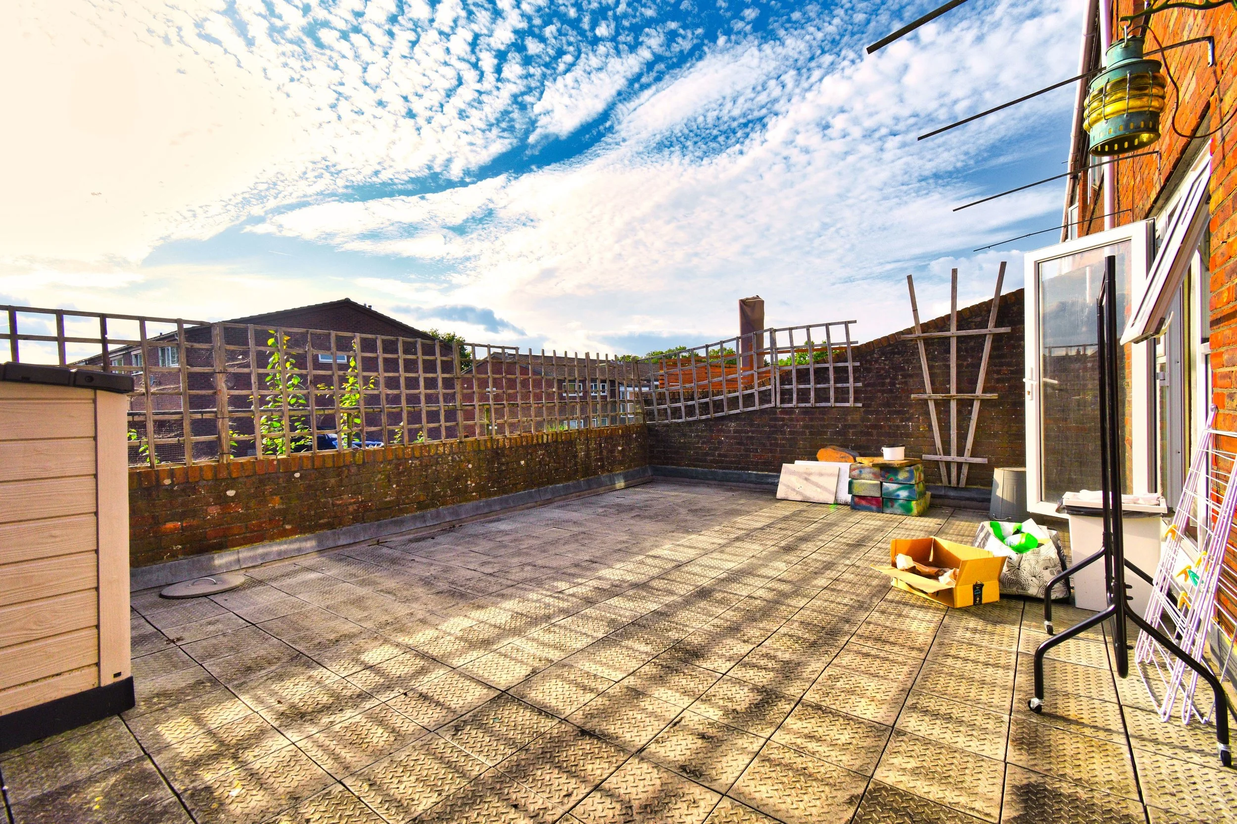 A rooftop terrace area with tiled flooring, brick walls, a view of neighboring houses, and a partly cloudy sky. There are some gardening trellises, boxes, and miscellaneous items, including a white door with glass and some hooks on the wall.