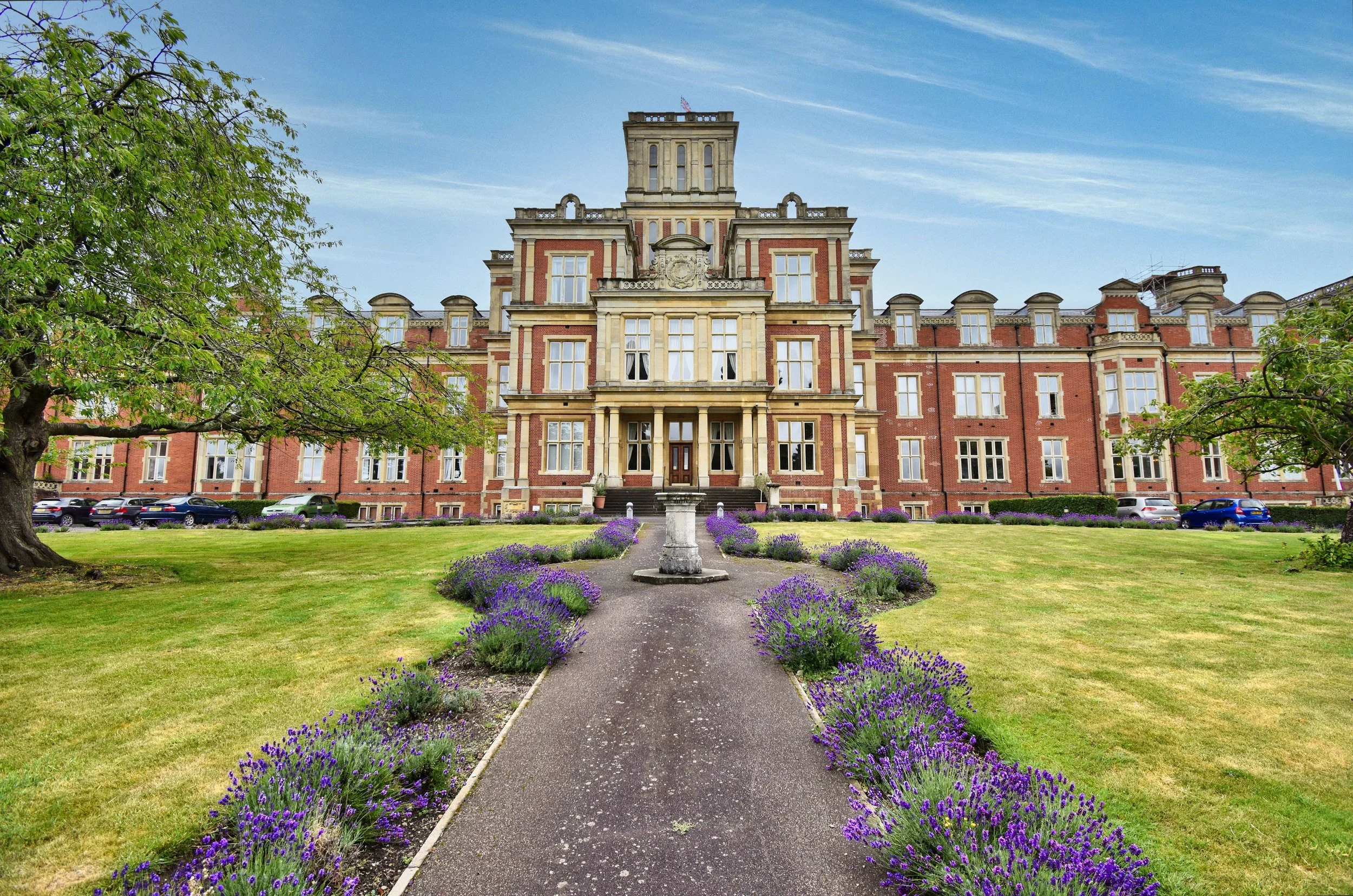 A large, ornate red brick and stone mansion with multiple stories, surrounded by a well-maintained garden with purple flowers, green grass, and trees, under a partly cloudy sky.