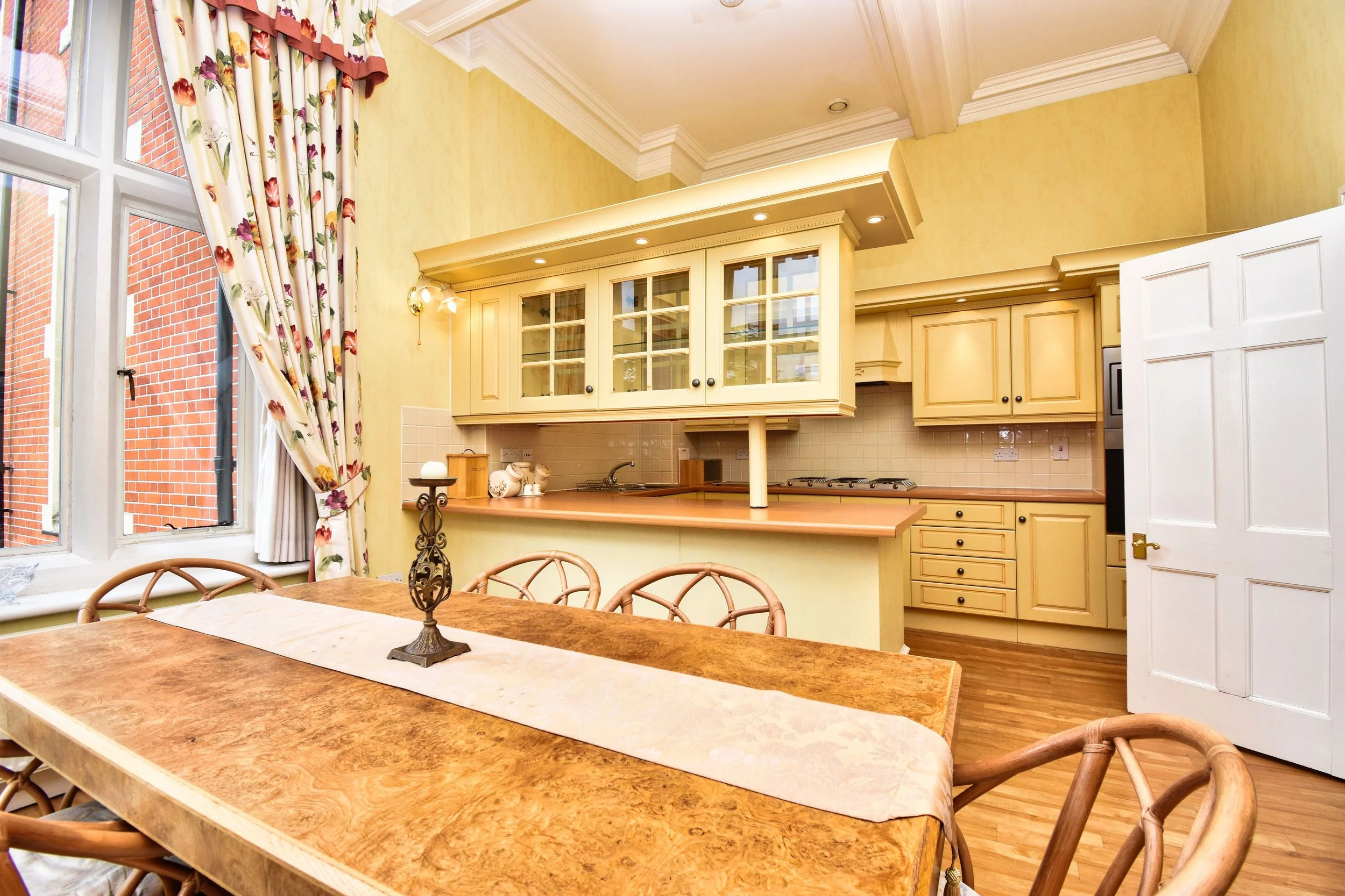 View of a kitchen with yellow cabinetry, a beige backsplash, and a wooden table with chairs. Large window with floral curtains shows a brick building outside.