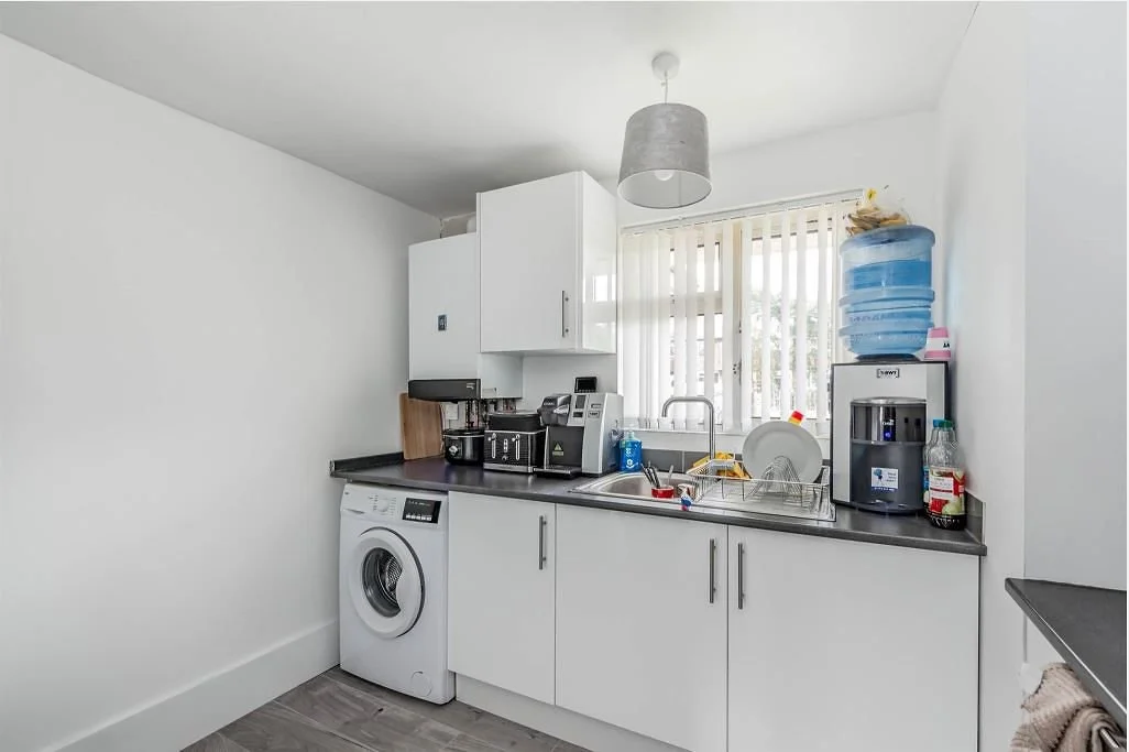 Kitchen with white cabinets, a black countertop, a washing machine, a sink, and various appliances including a coffee machine, kettle, and blender. There is a window with vertical blinds and a water dispenser on the countertop.