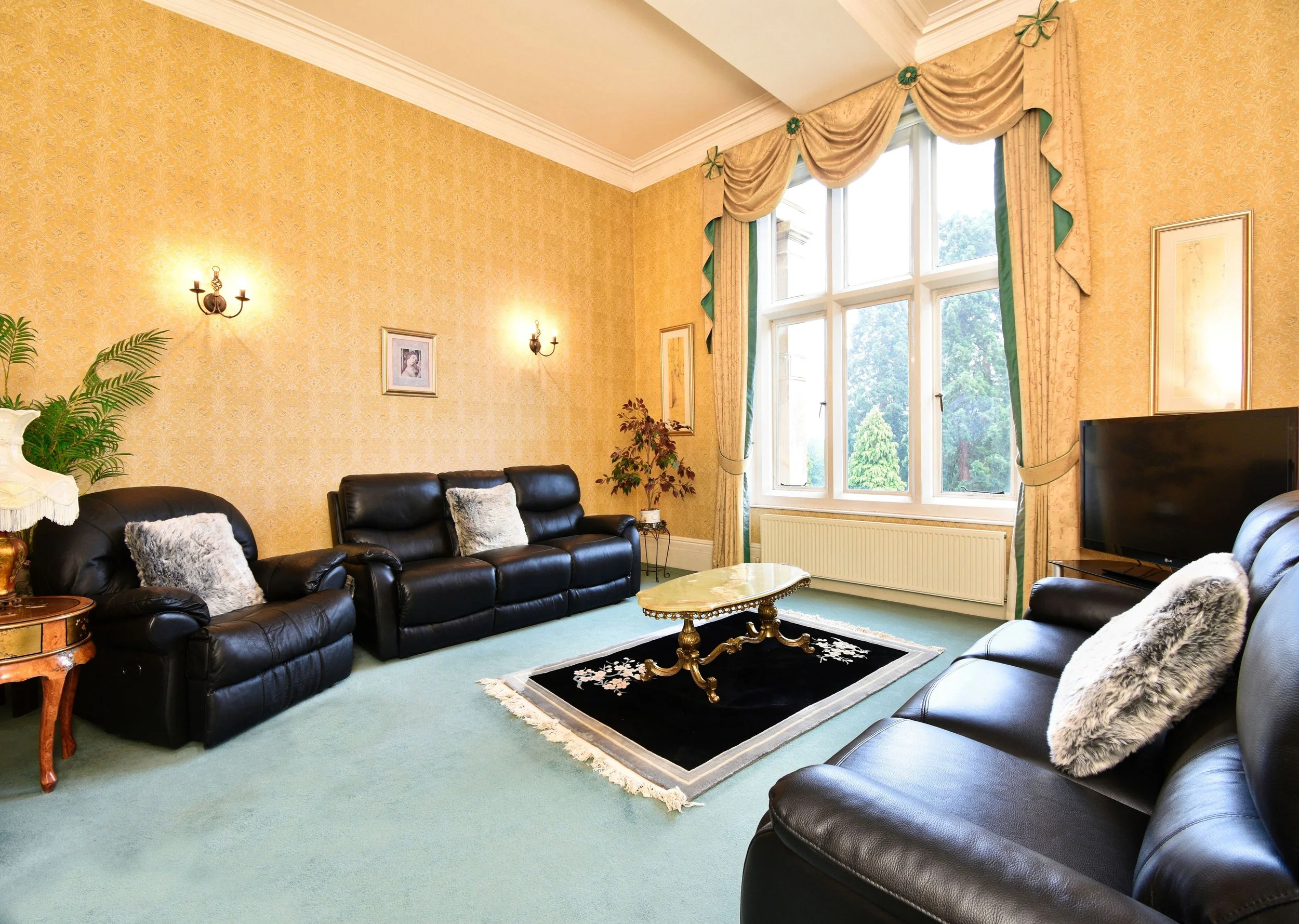 A living room with yellow patterned wallpaper, black leather sofas with fluffy pillows, a marble coffee table, framed artwork on the walls, large windows with gold and green curtains, a wall-mounted light fixture, a potted plant, and a small table.