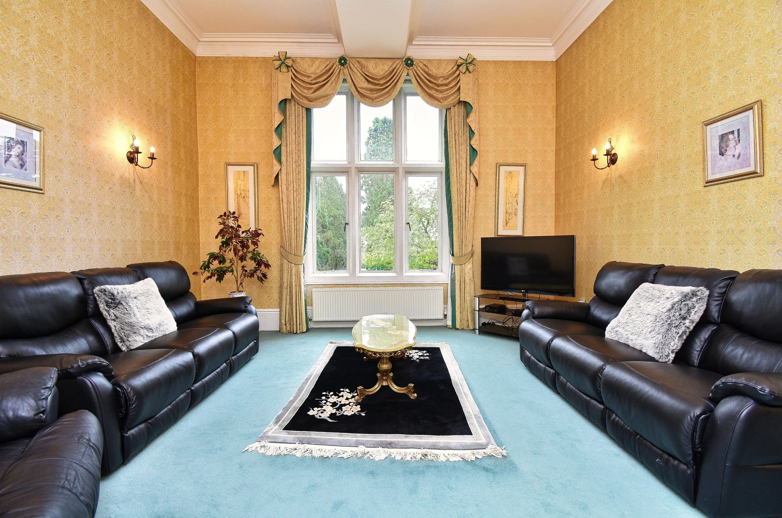 Living room with yellow wallpaper, dark leather sofas, a window with curtains, a small area rug, a TV, and decorative wall art