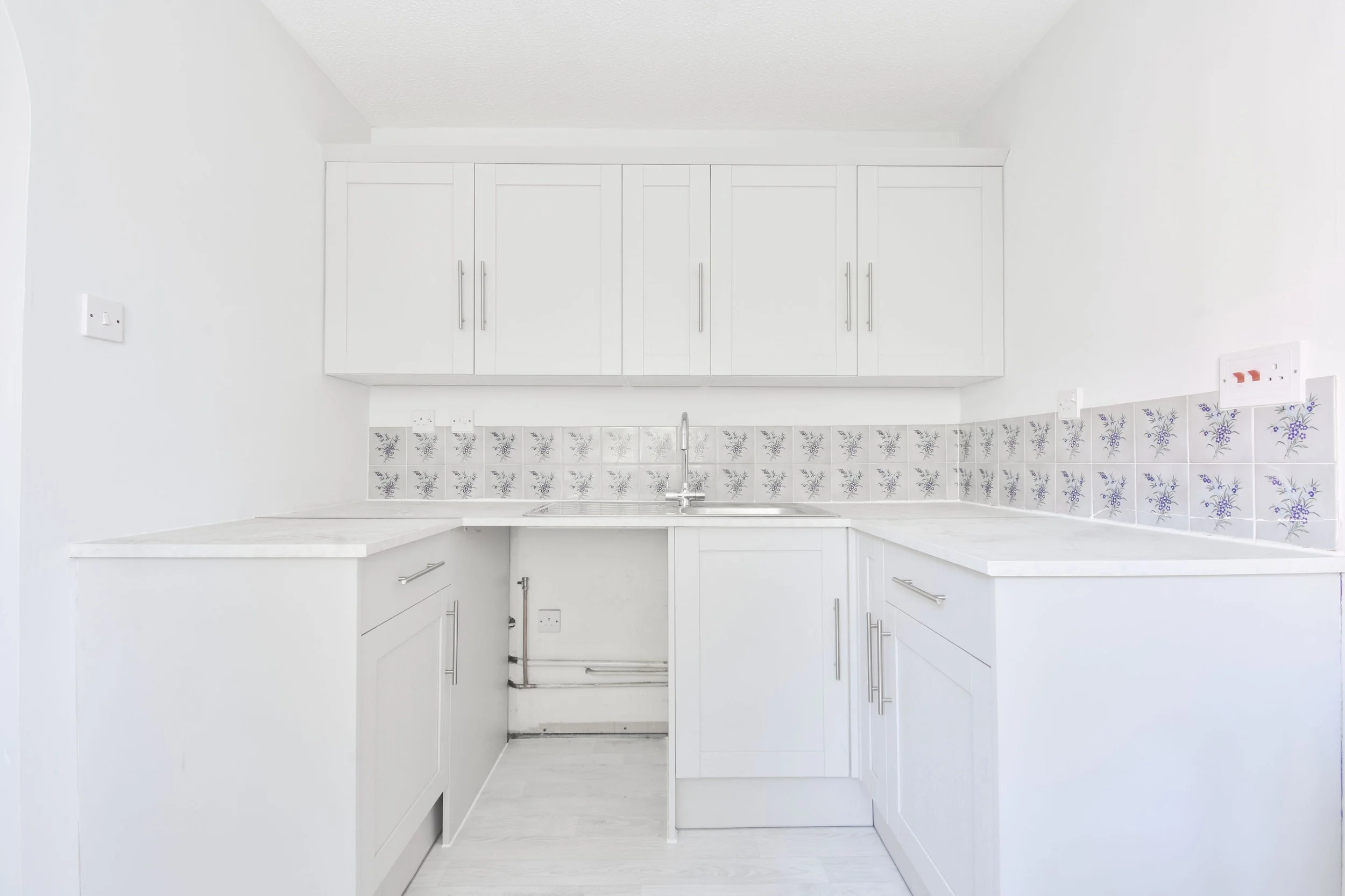 A white kitchen with white cabinets, a tiled backsplash with purple floral patterns, and a sink area with a faucet.