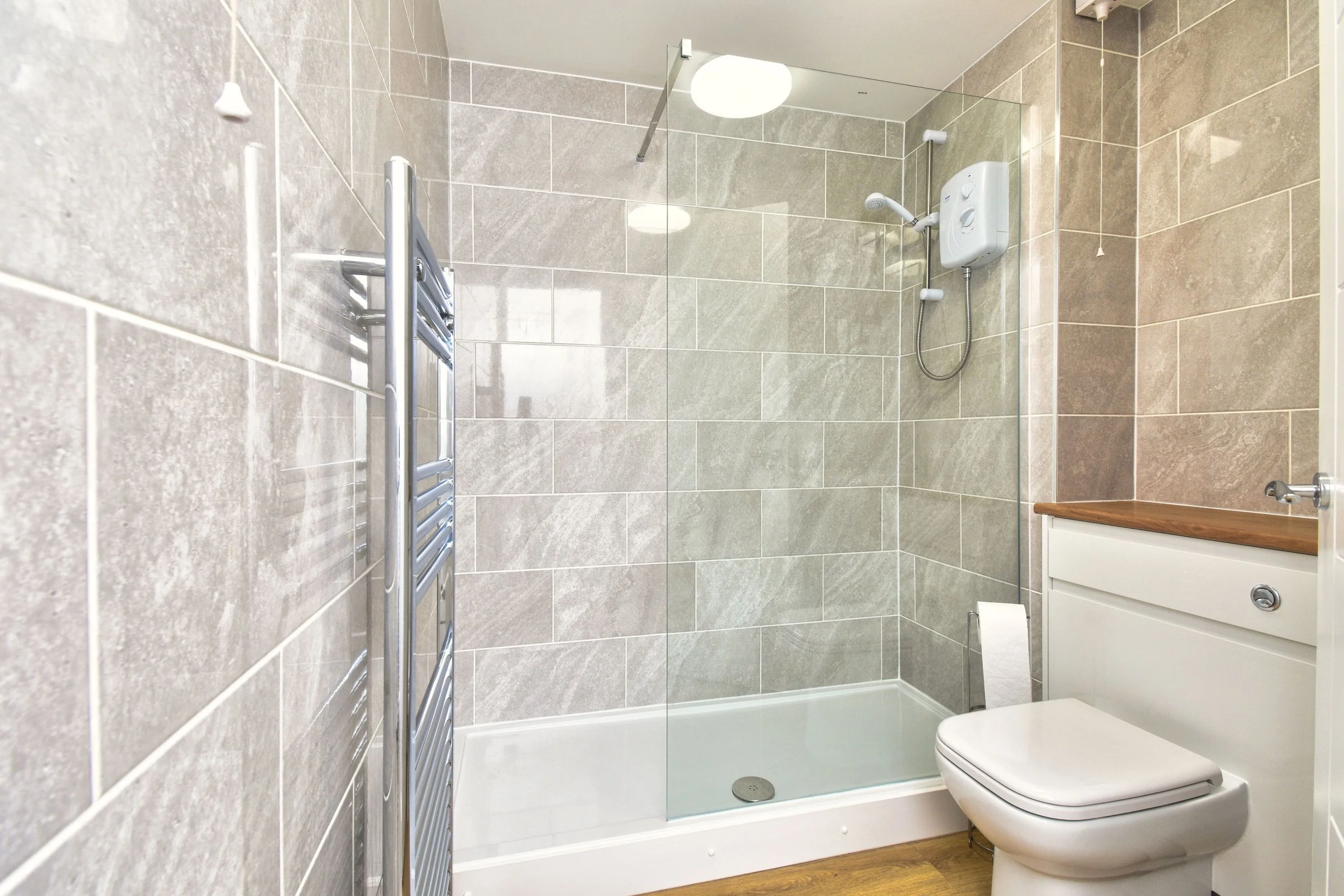 A modern bathroom with beige tiled walls, a walk-in shower with a glass partition, a stainless steel towel rack, a white electric shower unit, a small wooden cabinet with a white drawer, and a toilet. The flooring appears to be wooden.