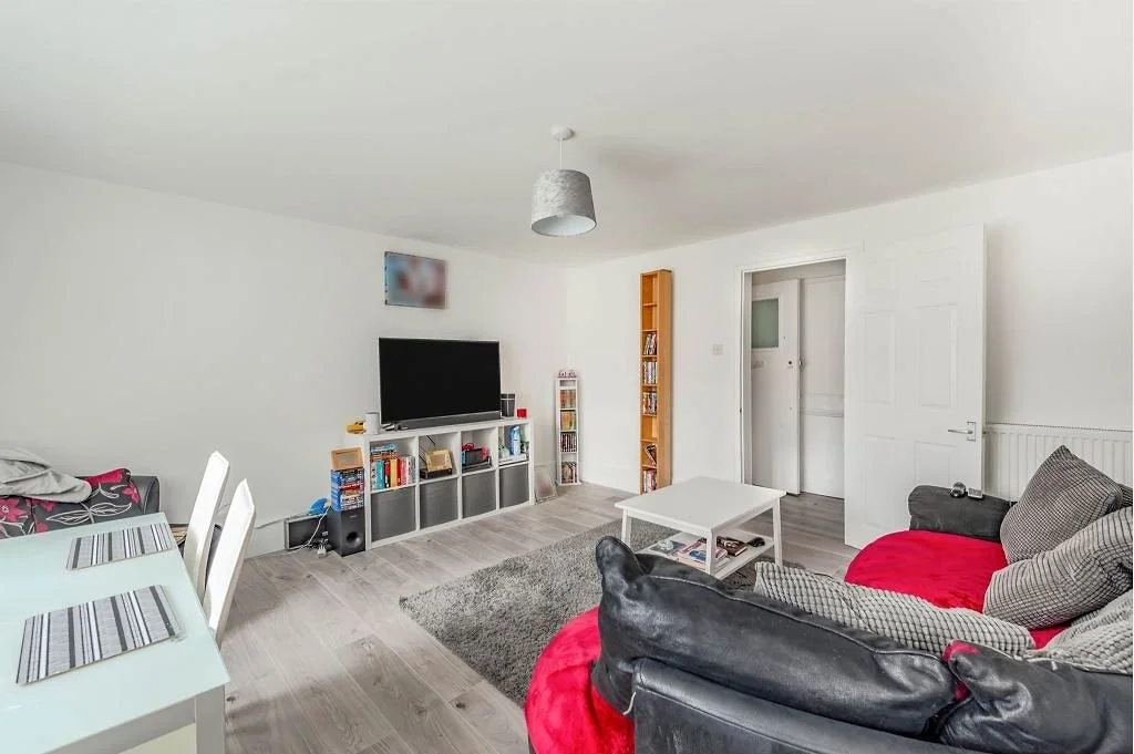 Living room with a white dining table and white chairs on the left, a black sofa with red and patterned pillows on the right, a white coffee table in the center, a gray rug, and a TV on a stand against the wall. There's a tall shelving unit next to t