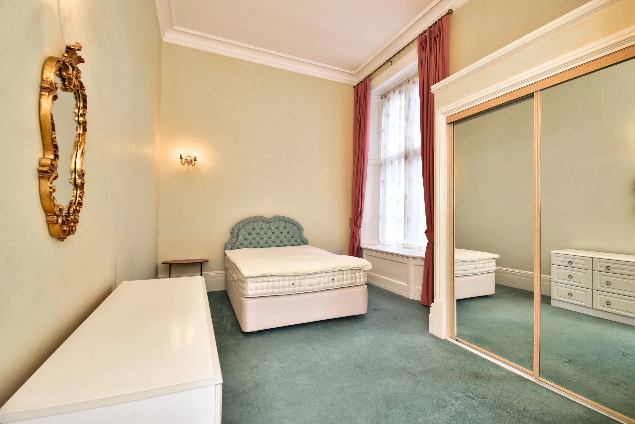A bedroom with a light green wall, a bed with a tufted headboard, a window with pink curtains, a mirror, a light fixture, and a white dresser.