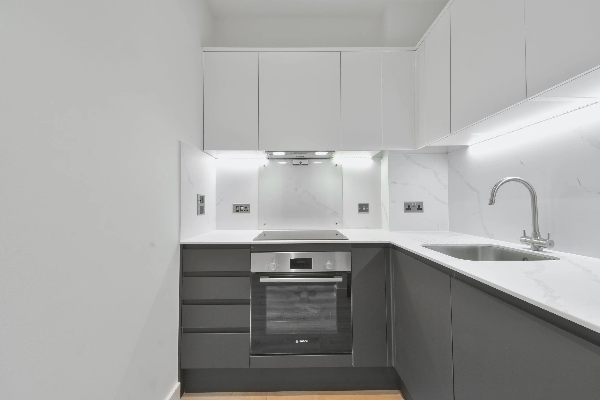 Modern kitchen with white walls, white marble backsplash, gray lower cabinets, and a stainless steel oven and sink. Overhead cabinets are white, and there is a built-in range hood with lighting.