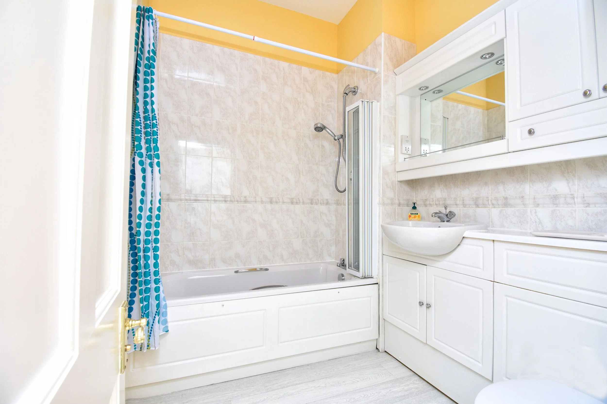 A small bathroom with a bathtub and shower combo, a sink, and white cabinetry. The walls are tiled with beige tiles, and the ceiling is painted yellow. There is a blue and white polka dot shower curtain.