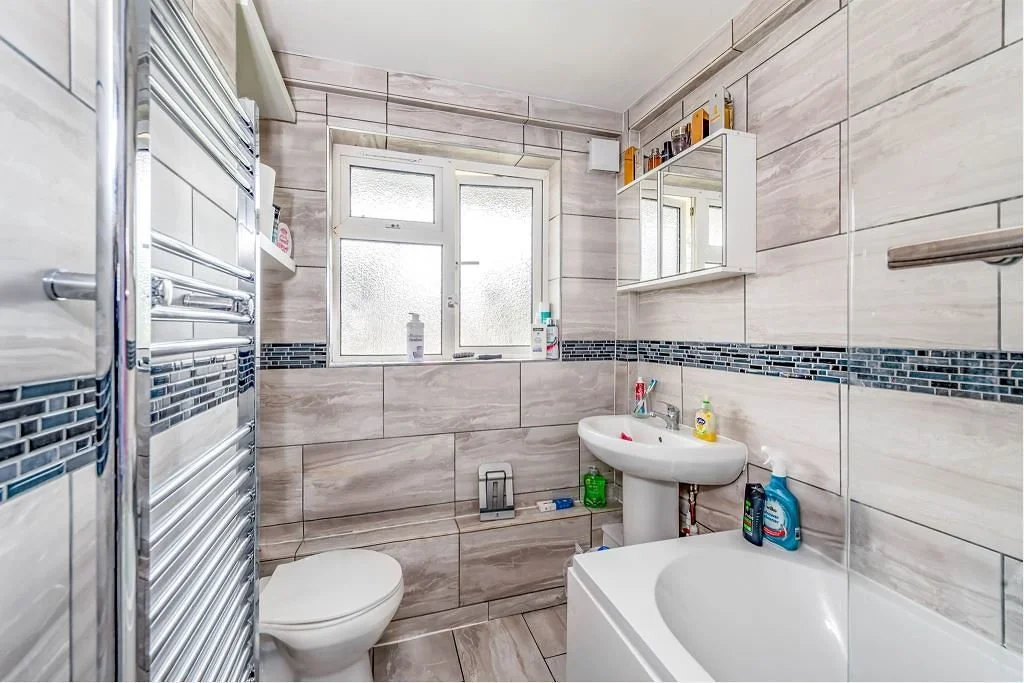 A modern small bathroom with beige tiled walls and floor, featuring a window, a toilet, a sink with toiletries, a bathtub, and a mirror cabinet.