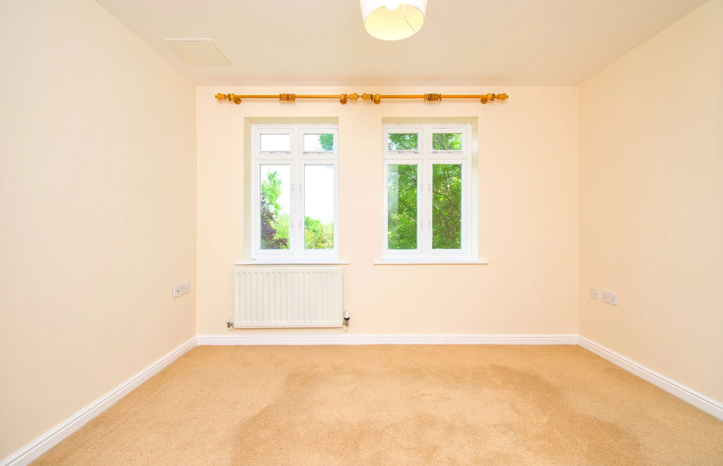 An empty beige carpeted room with off-white walls, a white radiator beneath a double-paned window, and a ceiling-mounted light fixture.