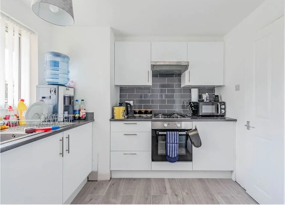 Minimalist white kitchen with gray countertops, gray tile backsplash, and various appliances including a microwave, toaster, and water dispenser. Dishes and cleaning supplies are on the counter, and a water bottle is placed on top of a water cooler.