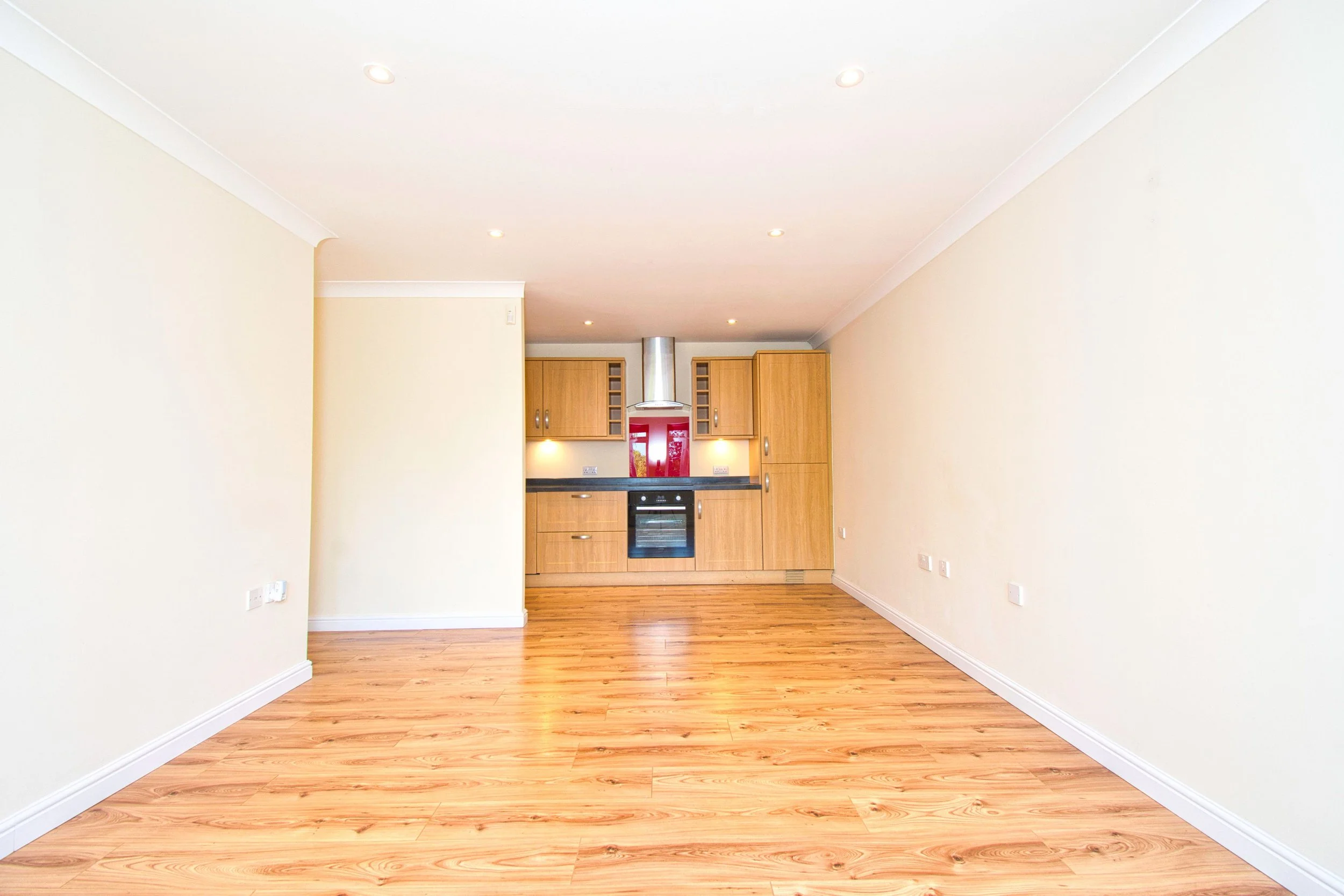 Empty living room with wood flooring, beige walls, and a view of a kitchen with wooden cabinets in the background.
