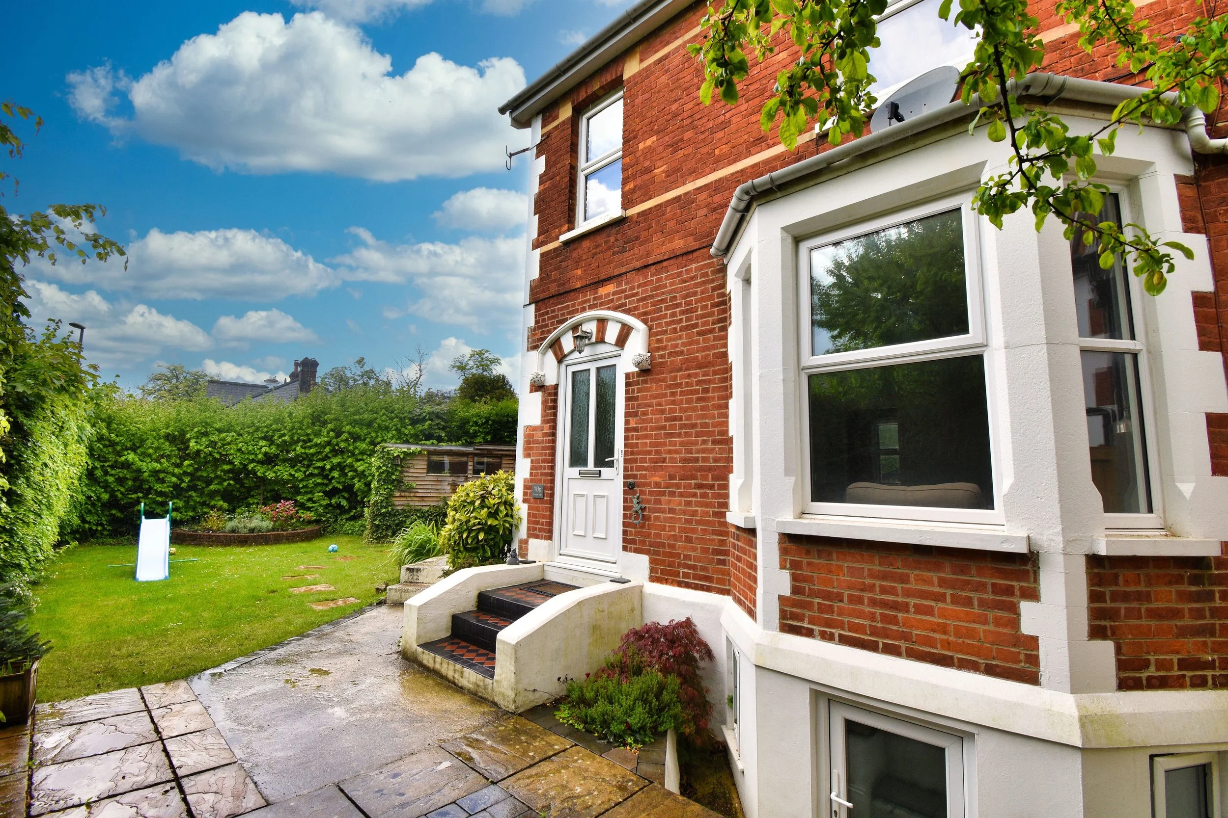 A red brick house with white trims and a bay window, surrounded by a lush green garden with a small slide, trees, and a cloudy sky.