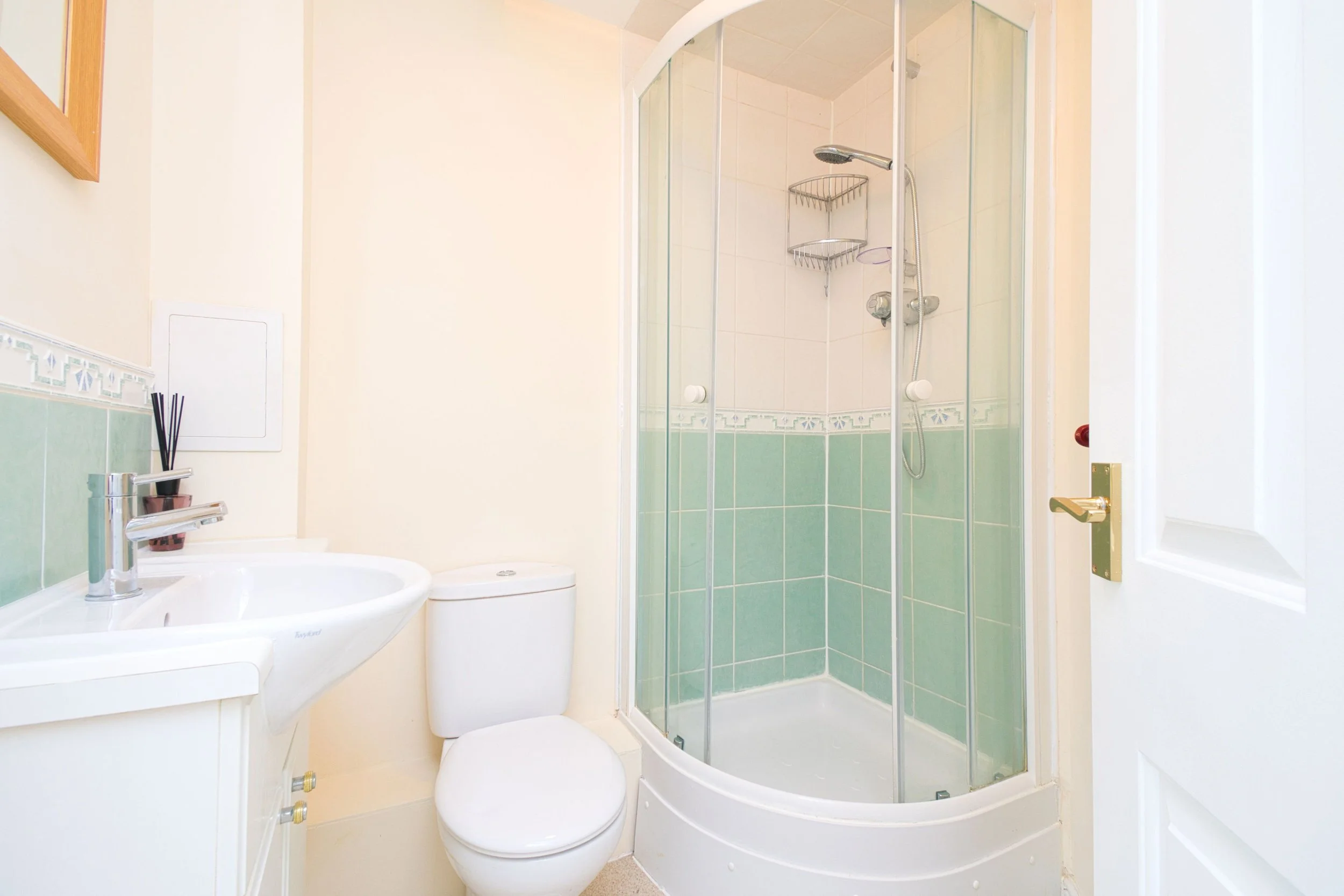 A small bathroom featuring a corner shower with clear glass doors, a white toilet, and a white sink with a faucet. The walls are partly tiled in green and beige, and there's a mirror and a small shelf inside the shower. A black reed diffuser is next 
