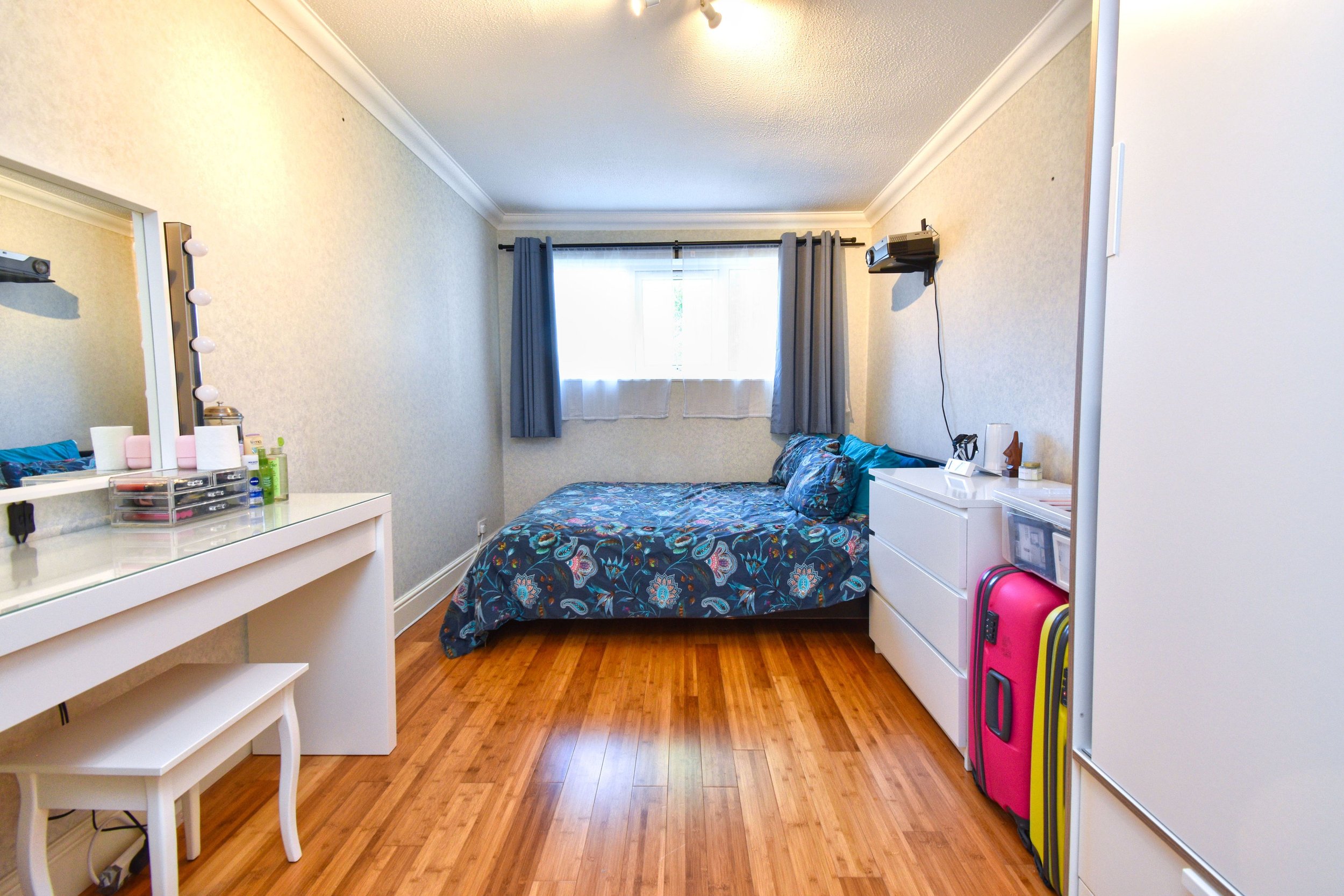 A bedroom with a bed against the back wall, a window with blue curtains, a white dresser with personal items, a pink suitcase, and a white vanity table with a mirror and makeup items.