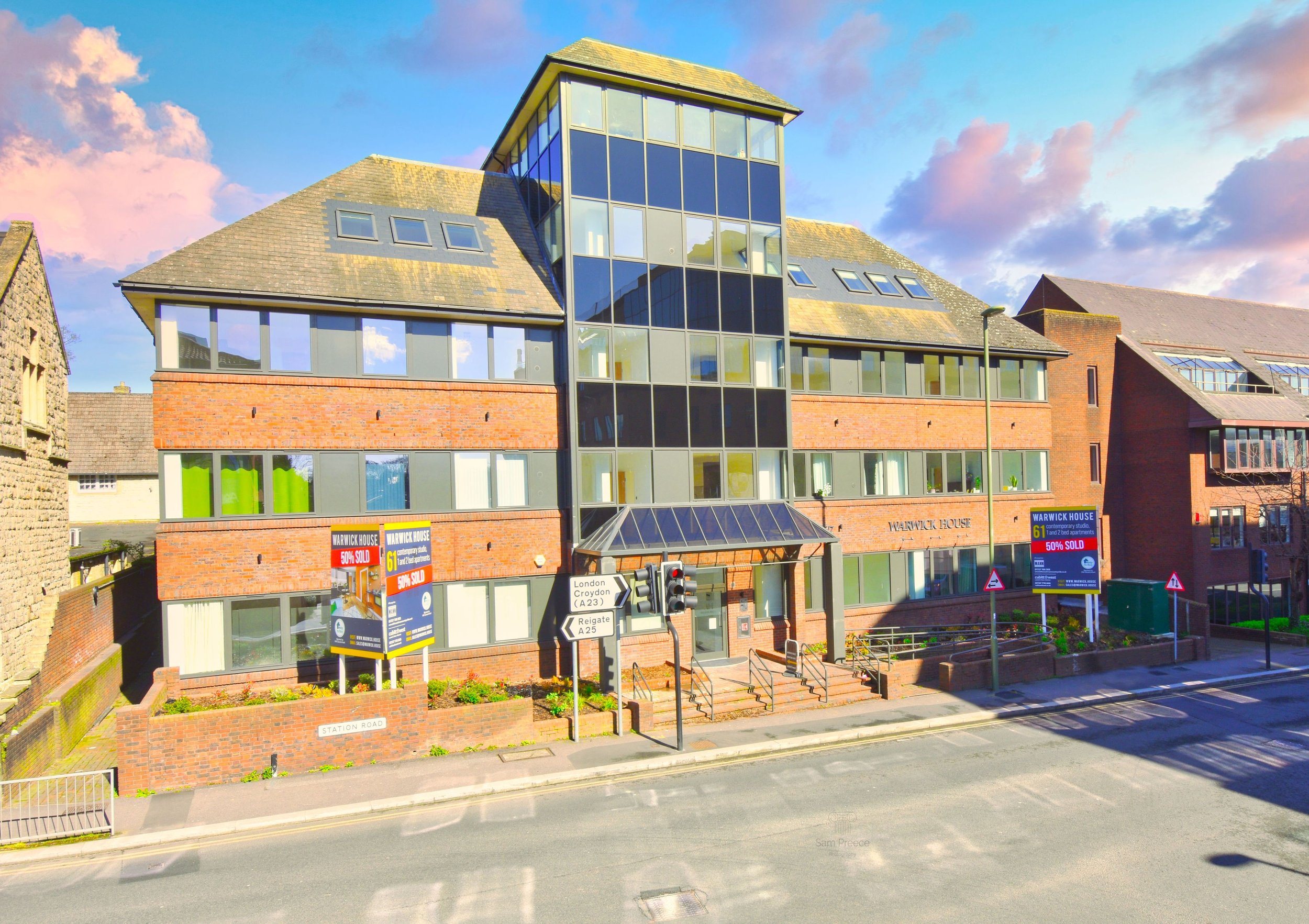 Modern multi-story building with brick and glass facade, with signs indicating 'Warwick House,' located on Station Road, near traffic signals and a street name sign. Bright blue sky with some clouds.
