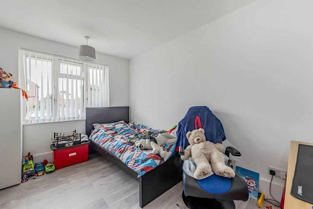 Child's bedroom with a bed, stuffed animals, toys, a chair with a teddy bear, and a window with vertical blinds.