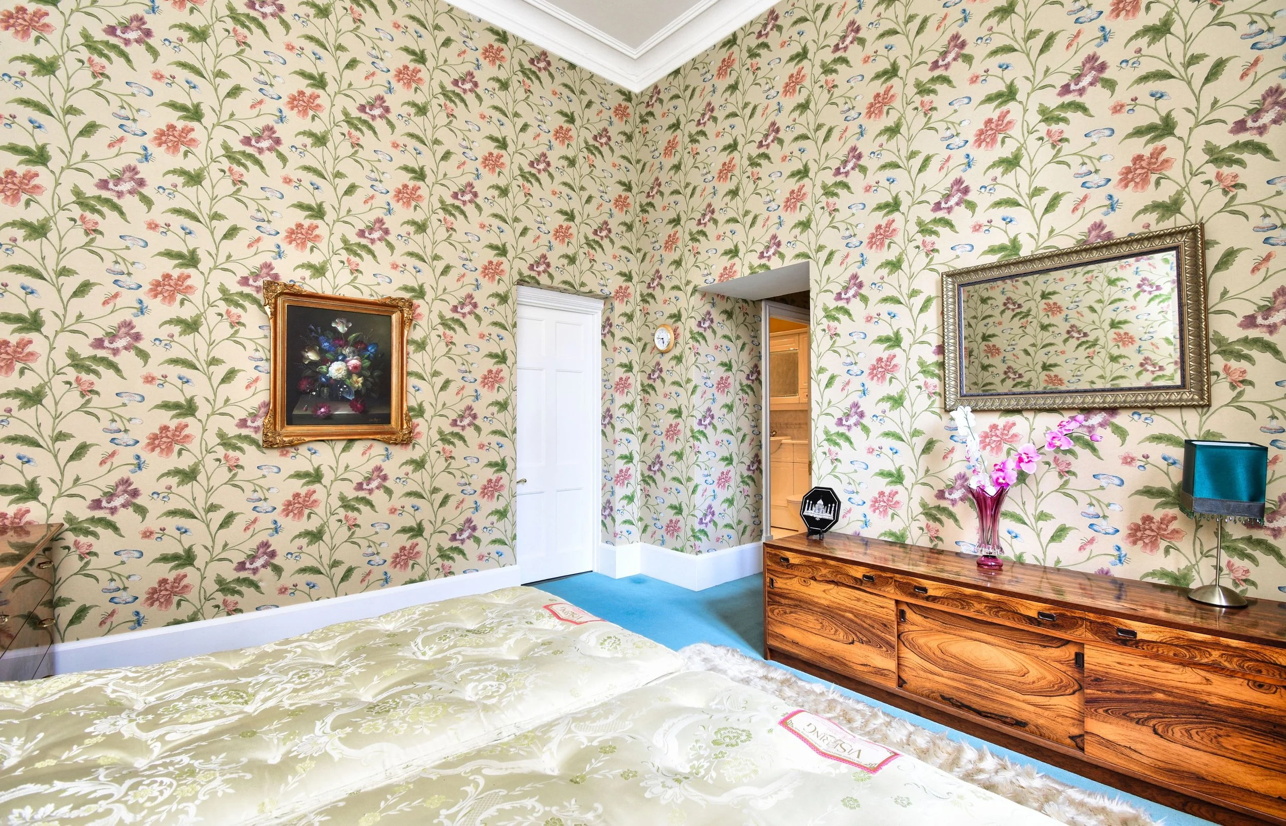 A bedroom with floral wallpaper, a wooden dresser, a mirror, paintings, a vase with pink flowers, and a green bedspread.