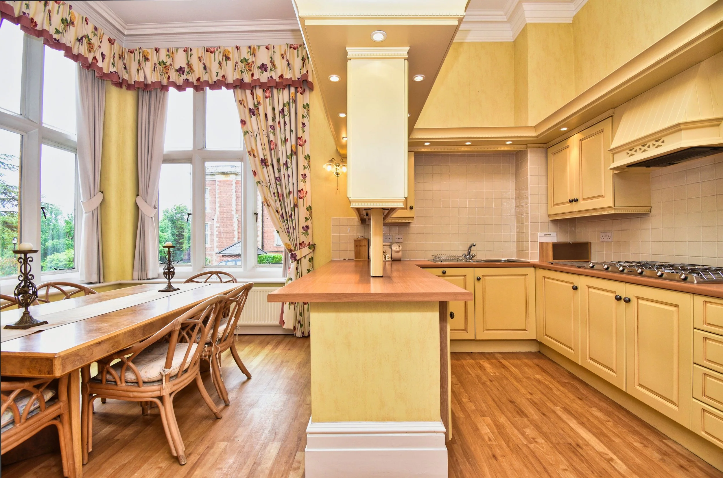 A kitchen and dining area with yellow cabinets, a wooden dining table with four chairs, a window with floral curtains, and a view of trees outside.