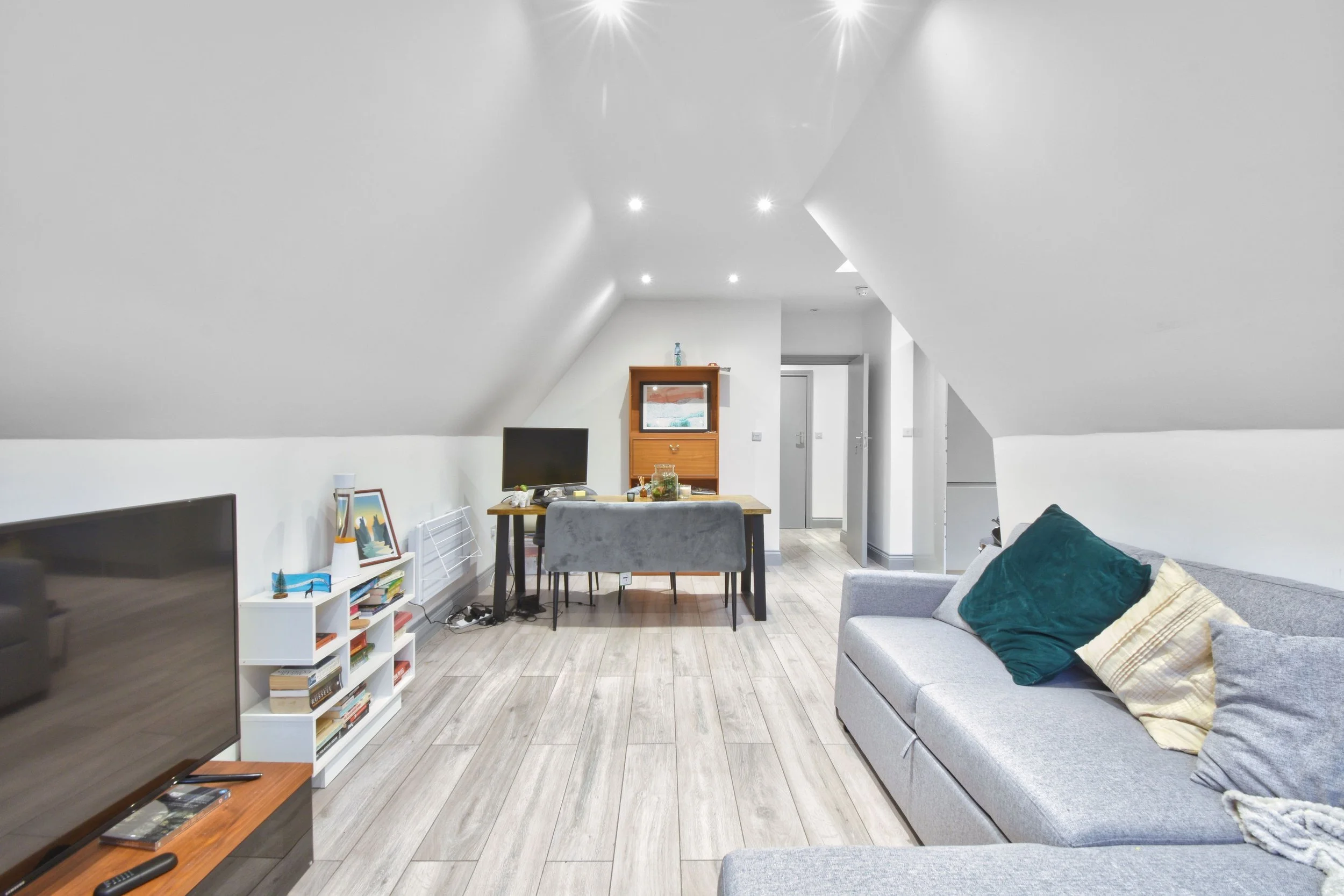 Living room with a gray sofa with pillows, a white bookshelf with books, a flat-screen TV, a wooden dining table with gray chairs, a small wooden cabinet, and a white door in the background, under a white sloped ceiling with recessed lighting.