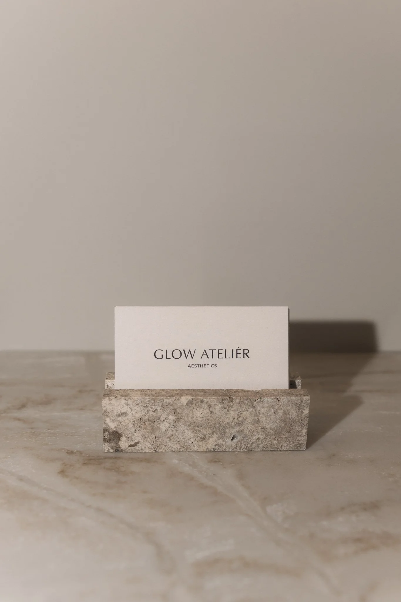 A small white card with the text 'GLOW ATELIER AESTHETICS' sitting on a textured, rectangular stone block on a marble surface with a plain, light-colored wall background.