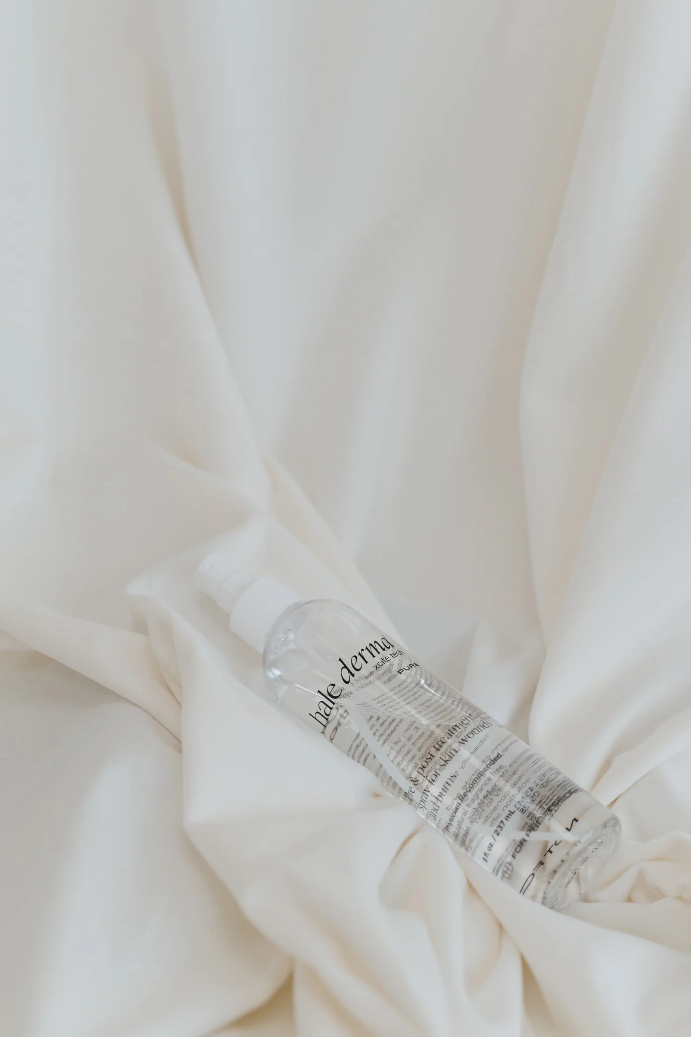 A transparent bottle of skincare serum lying on white fabric with folds and shadows.