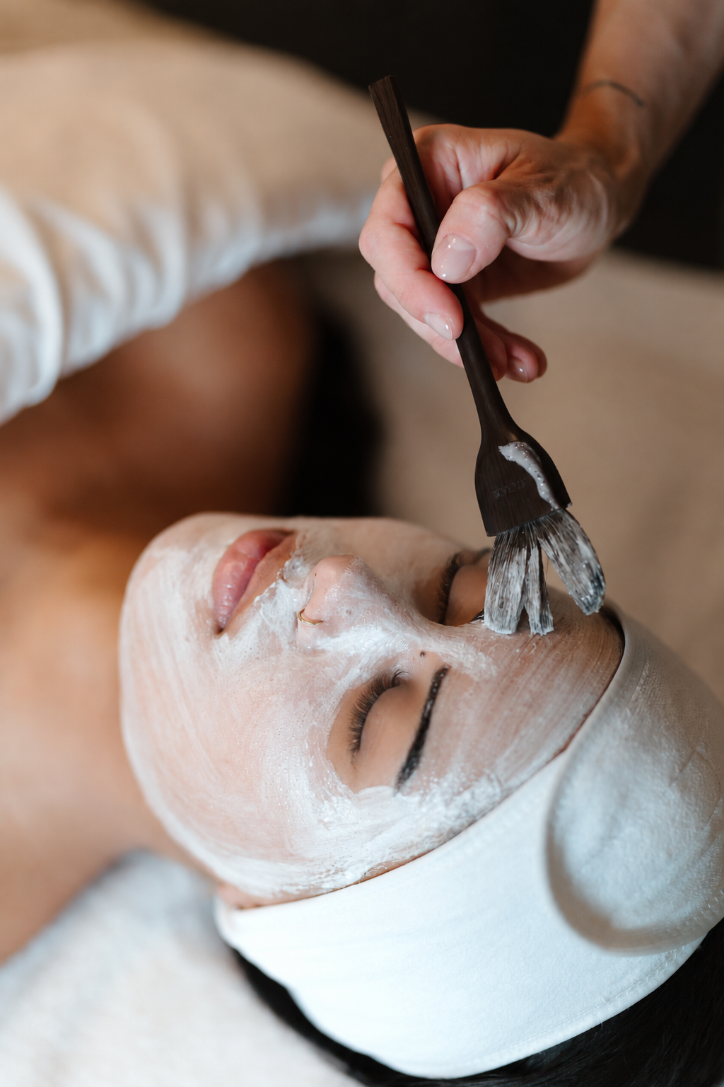 A woman receiving a facial treatment with a white mask applied on her face, lying down with eyes closed, while a technician brushes off excess mask with a small brush.