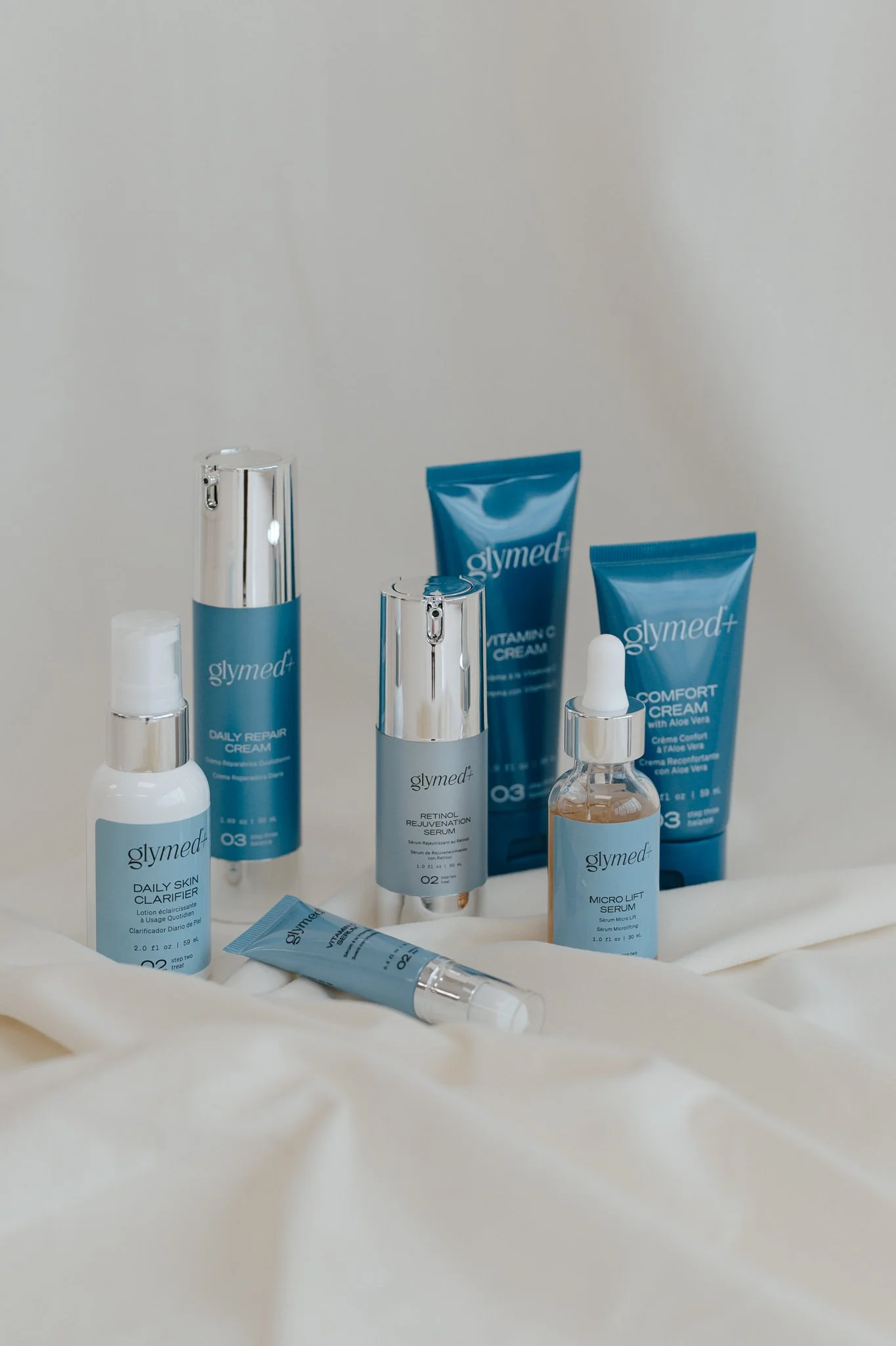 A collection of skincare products from the brand Glymed, including creams, serums, and a clarifier, arranged on a light-colored fabric surface.