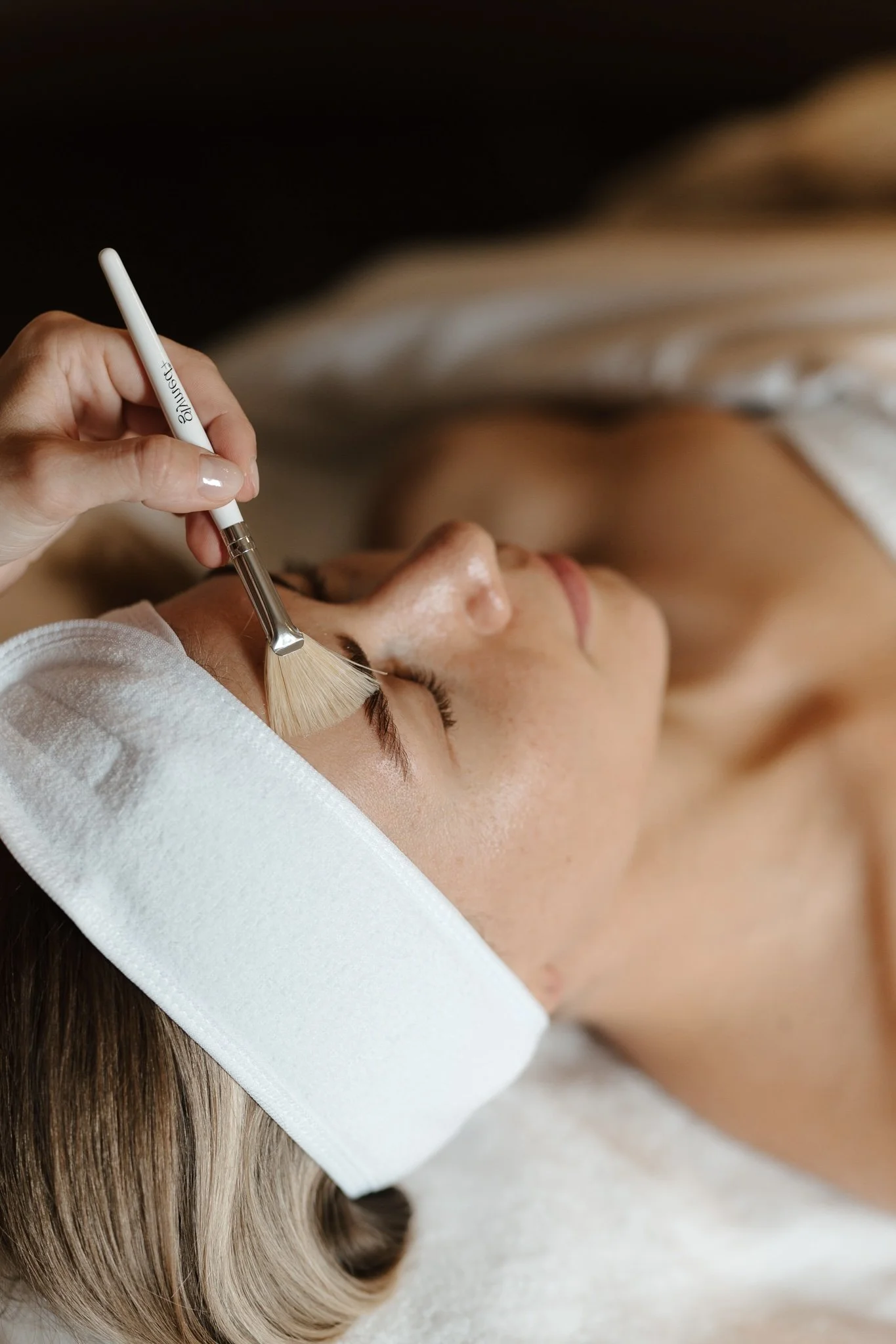 A woman receiving a facial treatment with a brush. The woman is lying down with her eyes closed, and the technician is applying product to her forehead.