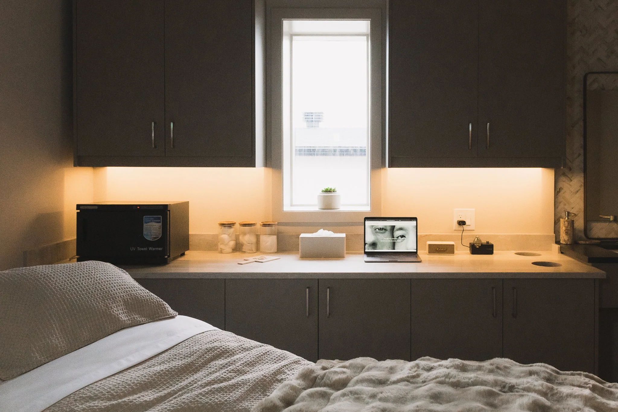 A modern bedroom desk area by the window with a small potted plant, jars, a laptop, a microwave, and other small items. Part of a bed with textured bedding is visible in the foreground.
