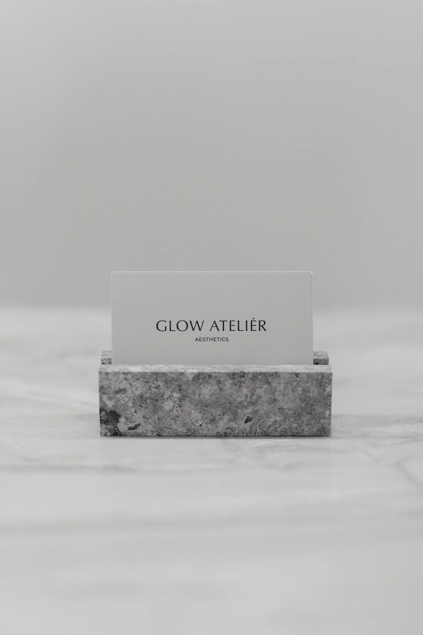 A white card with the text 'GLOW ATELIER' and 'AESTHETICS' sits on a textured stone block with a plain gray background.
